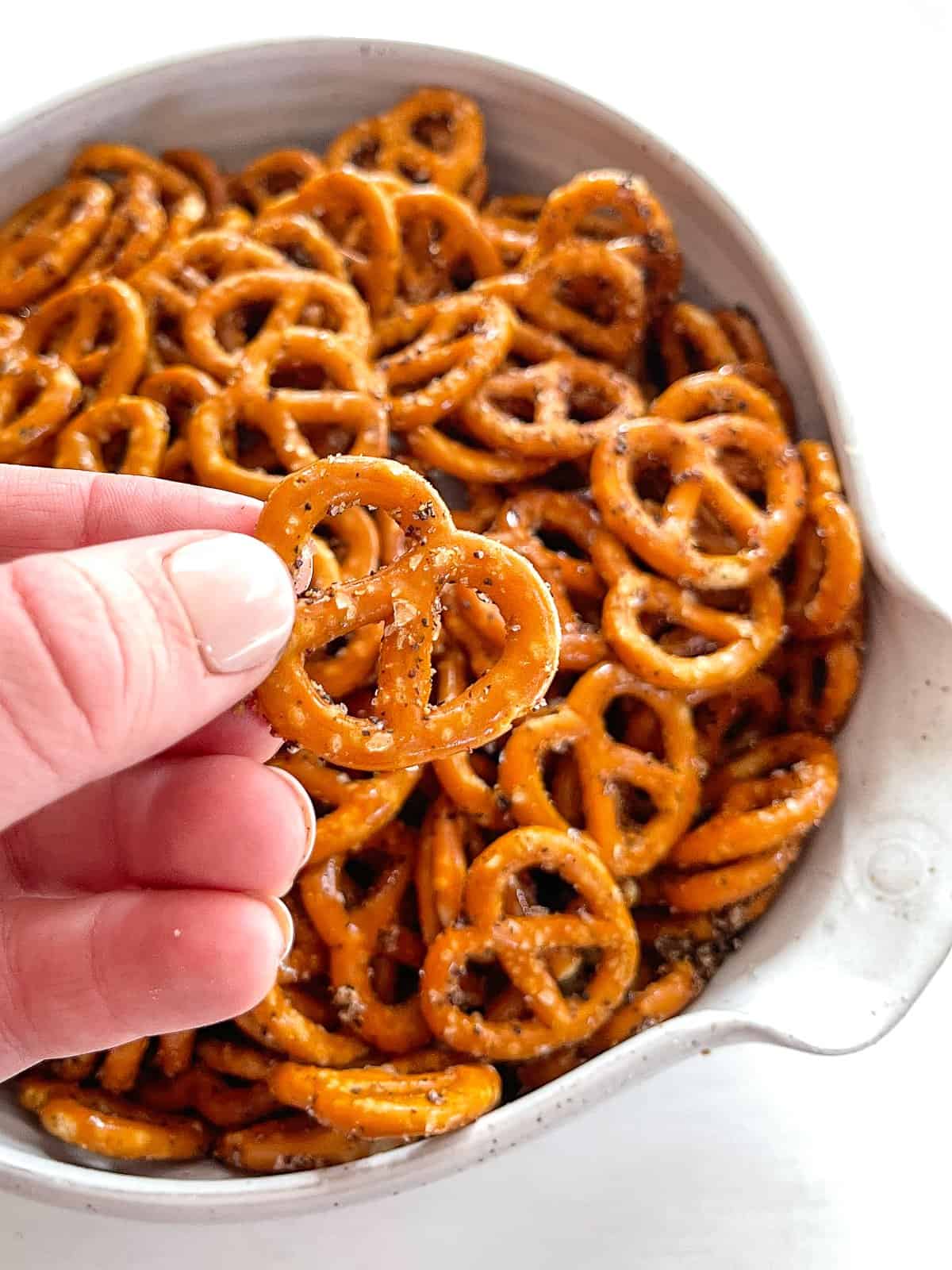 a close up photo of a white bowl filled with pretzel twists with a hand holding one pretzel close to the camera