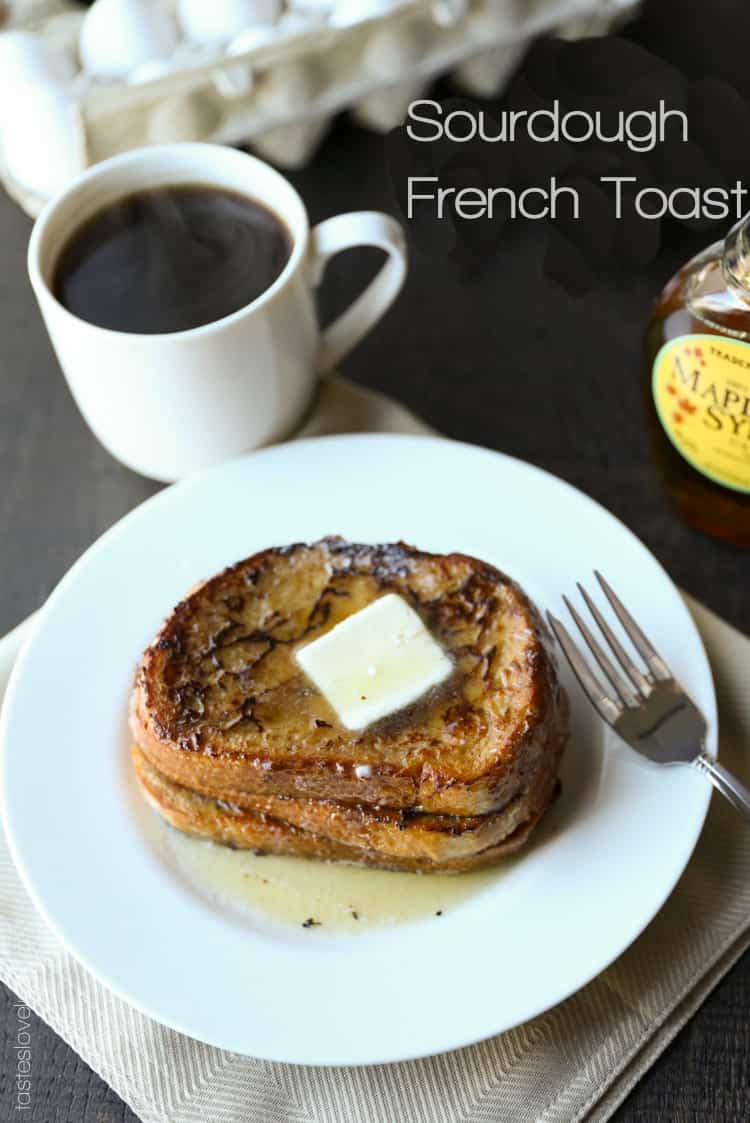 three slices of french toast on a white plate topped with a square of butter with a silver fork resting on the plate and a white mug of coffee and bottle of maple syrup in the background