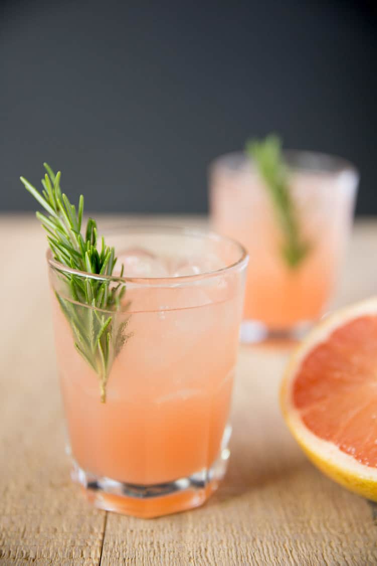 two glasses of rosemary greyhound cocktails garnished with a sprig of rosemary next to half a grapefruit