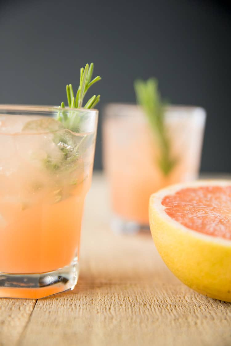 two glasses filled with rosemary greyhound cocktails next to half a grapefruit