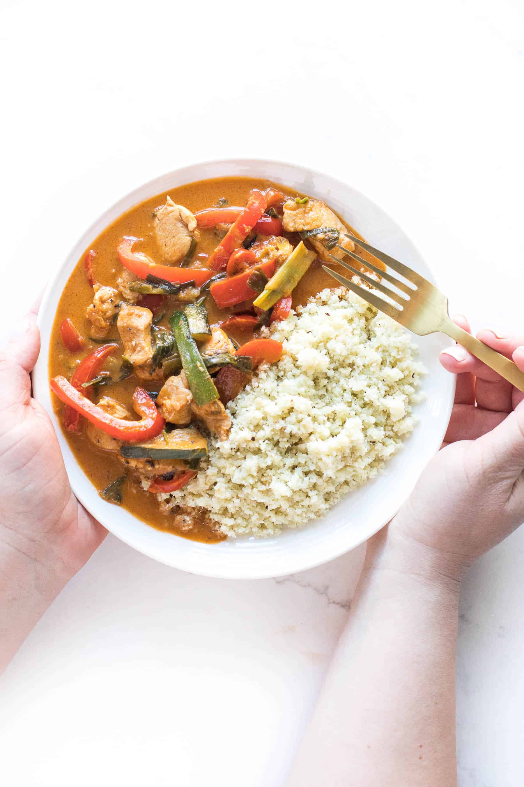 2 hands holding a white plate of chicken curry with low carb cauliflower rice