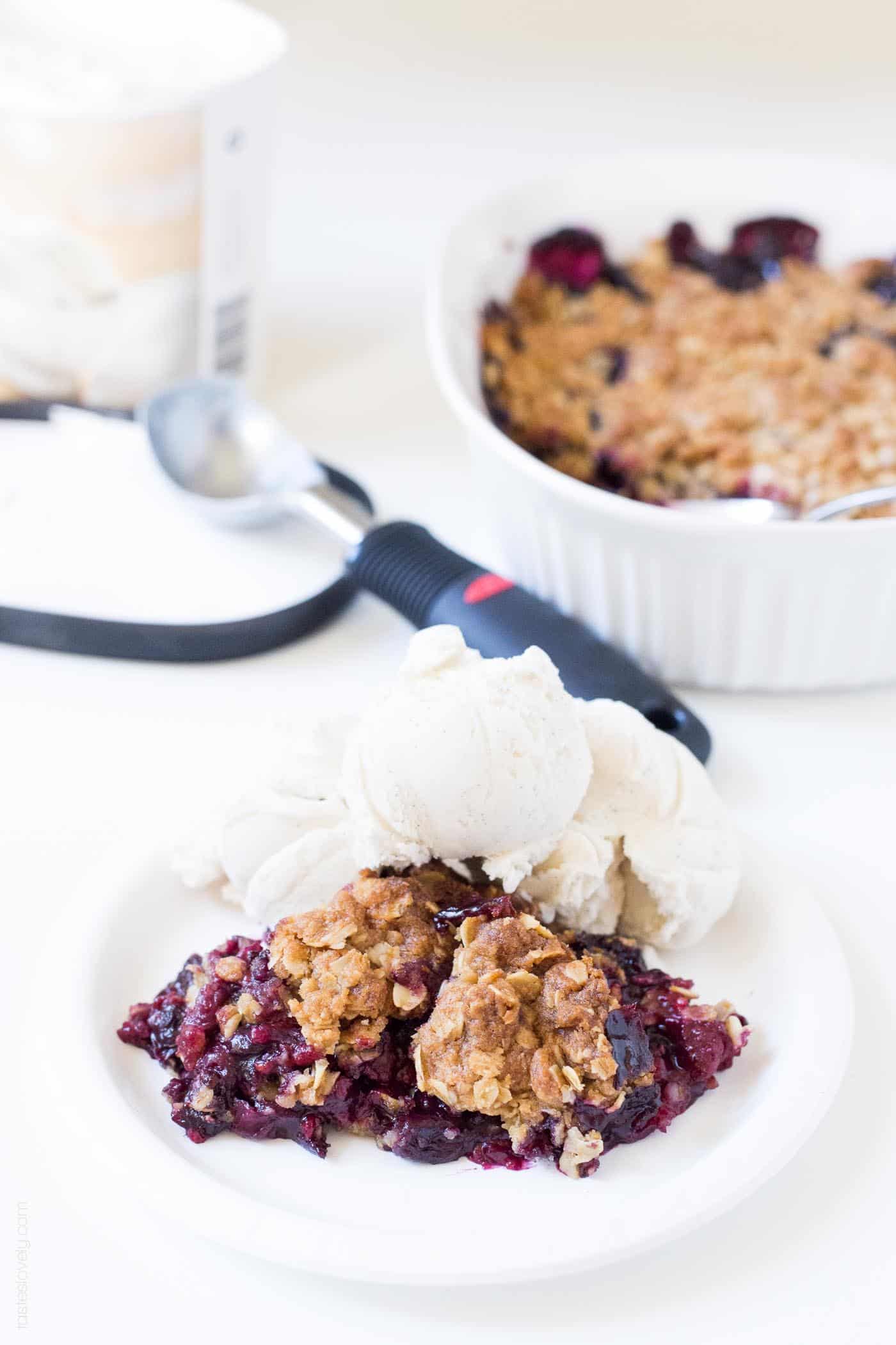 a white plate with a piece of triple berry crumble recipe and a scoop of vanilla ice cream with a black ice cream scooper in the background