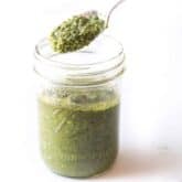 A spoon is lifting a portion of green pesto sauce from a glass jar. The jar, with Ball and Wide Mouth embossed on it, is filled with vibrant green pesto, sitting on a white surface.