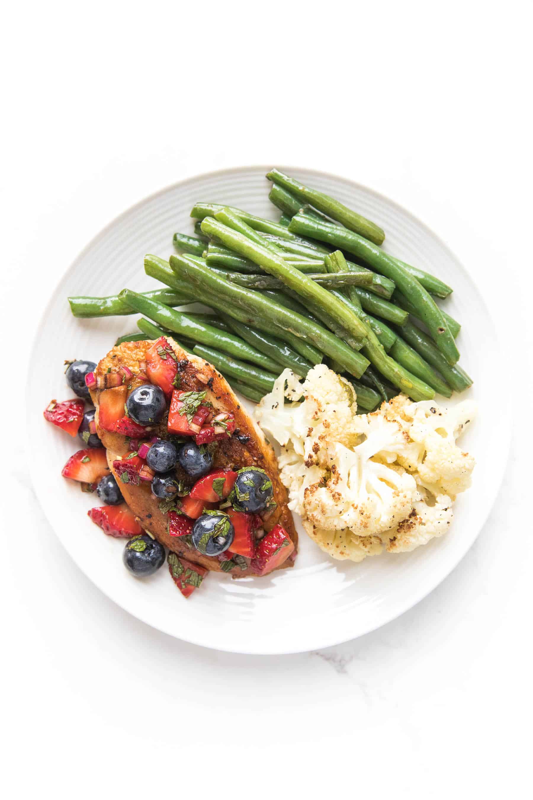 chicken topped with blueberry and strawberry salsa on a white plate and a white background with green beans and cauliflower