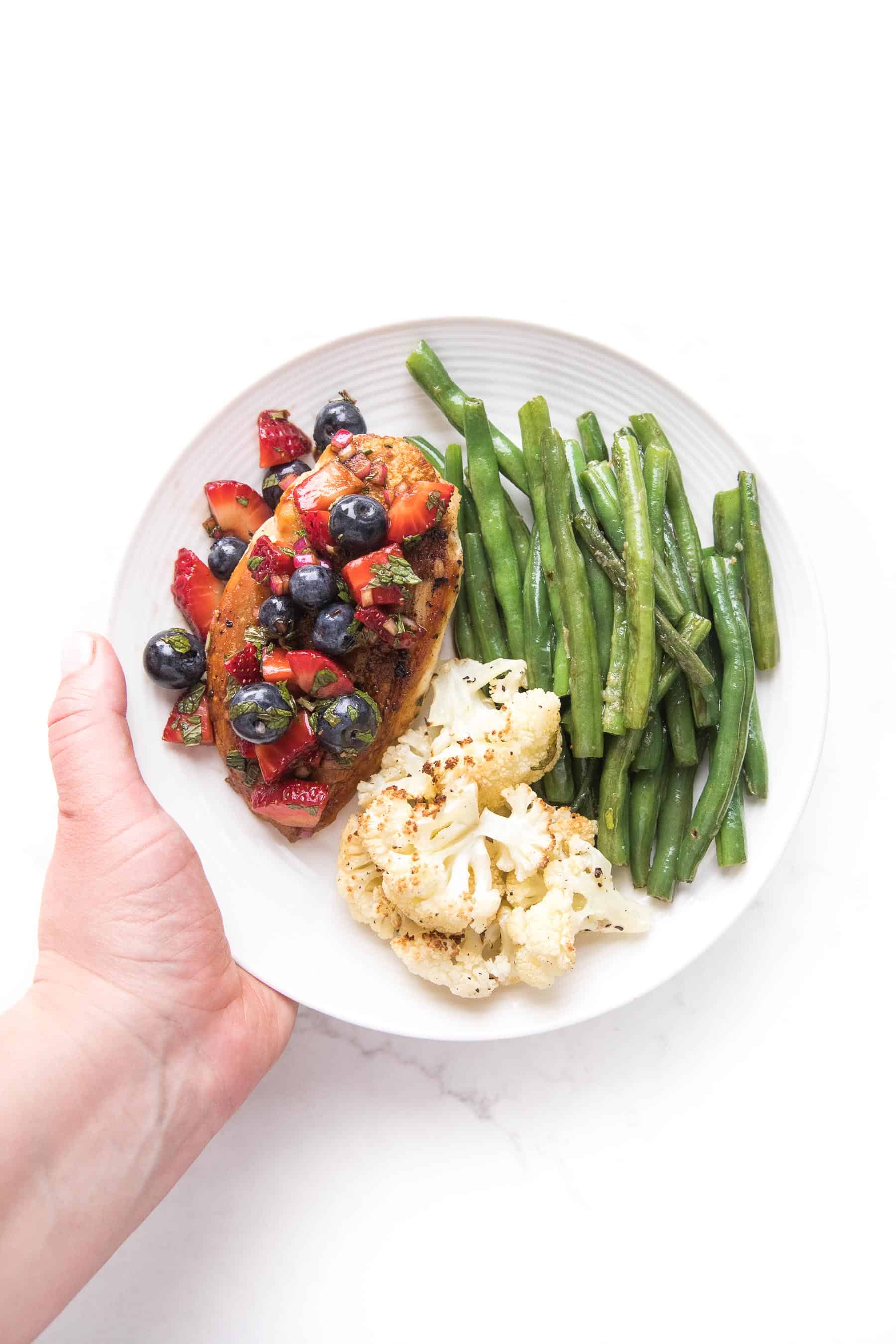 a hand holding chicken topped with blueberry and strawberry salsa on a white plate and a white background