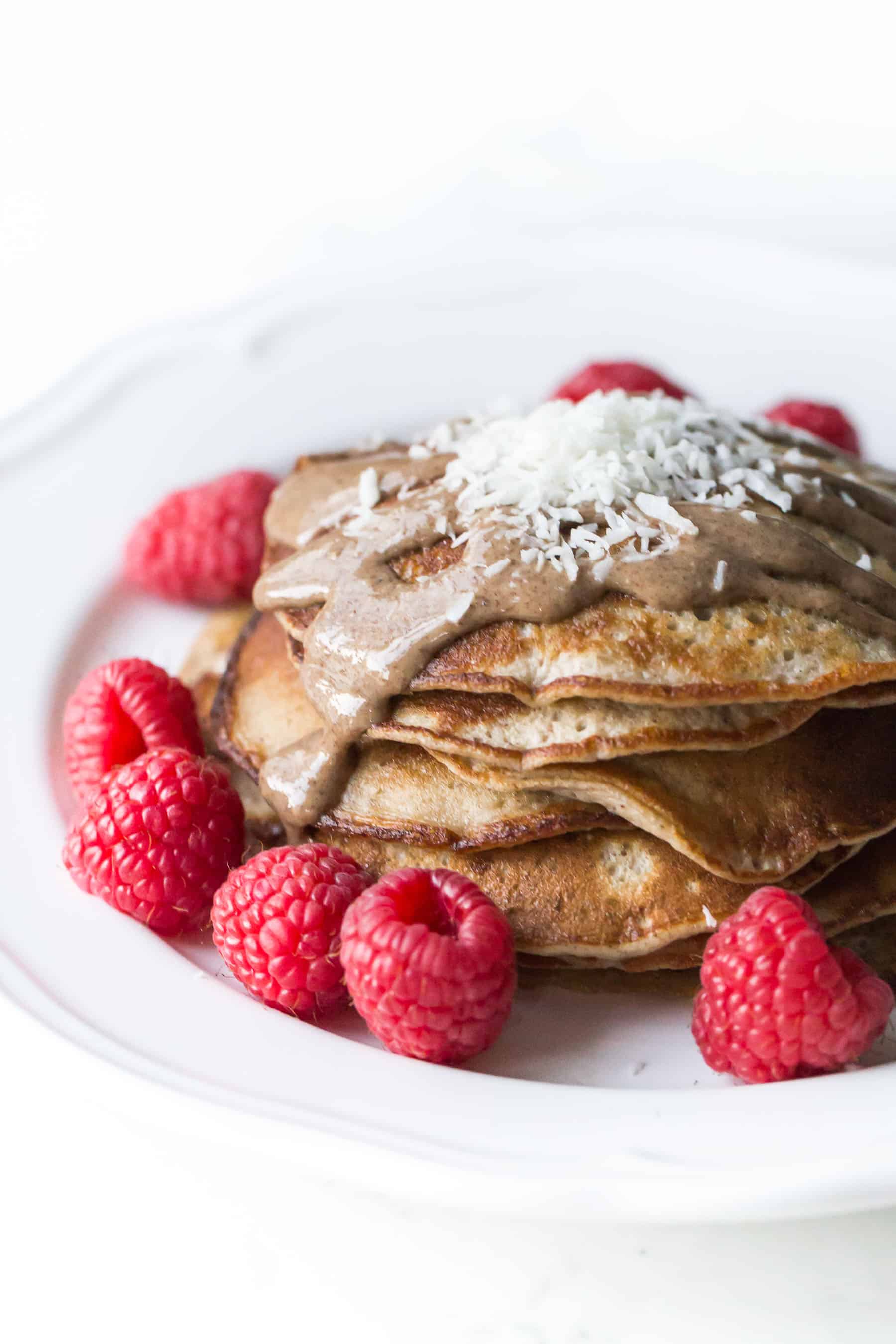 pancakes on a white plate topped with almond butter, shredded coconut and raspberries