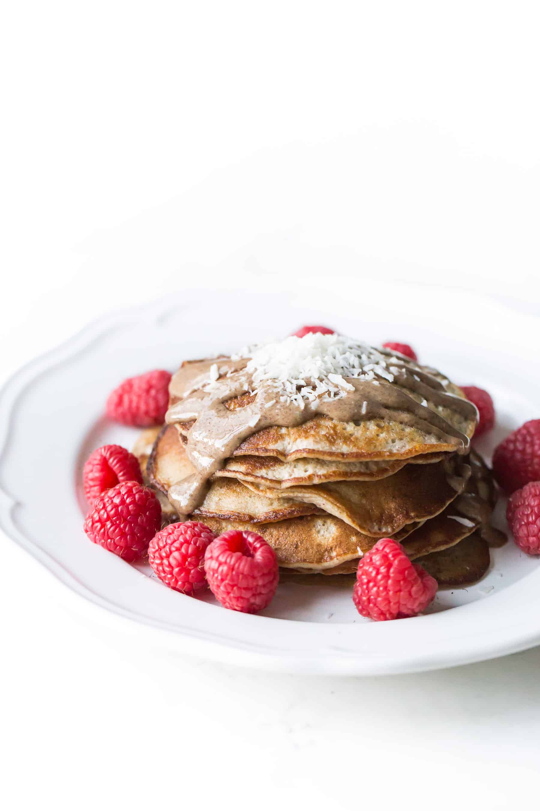 small pancakes on a white plate topped with almond butter, shredded coconut and raspberries