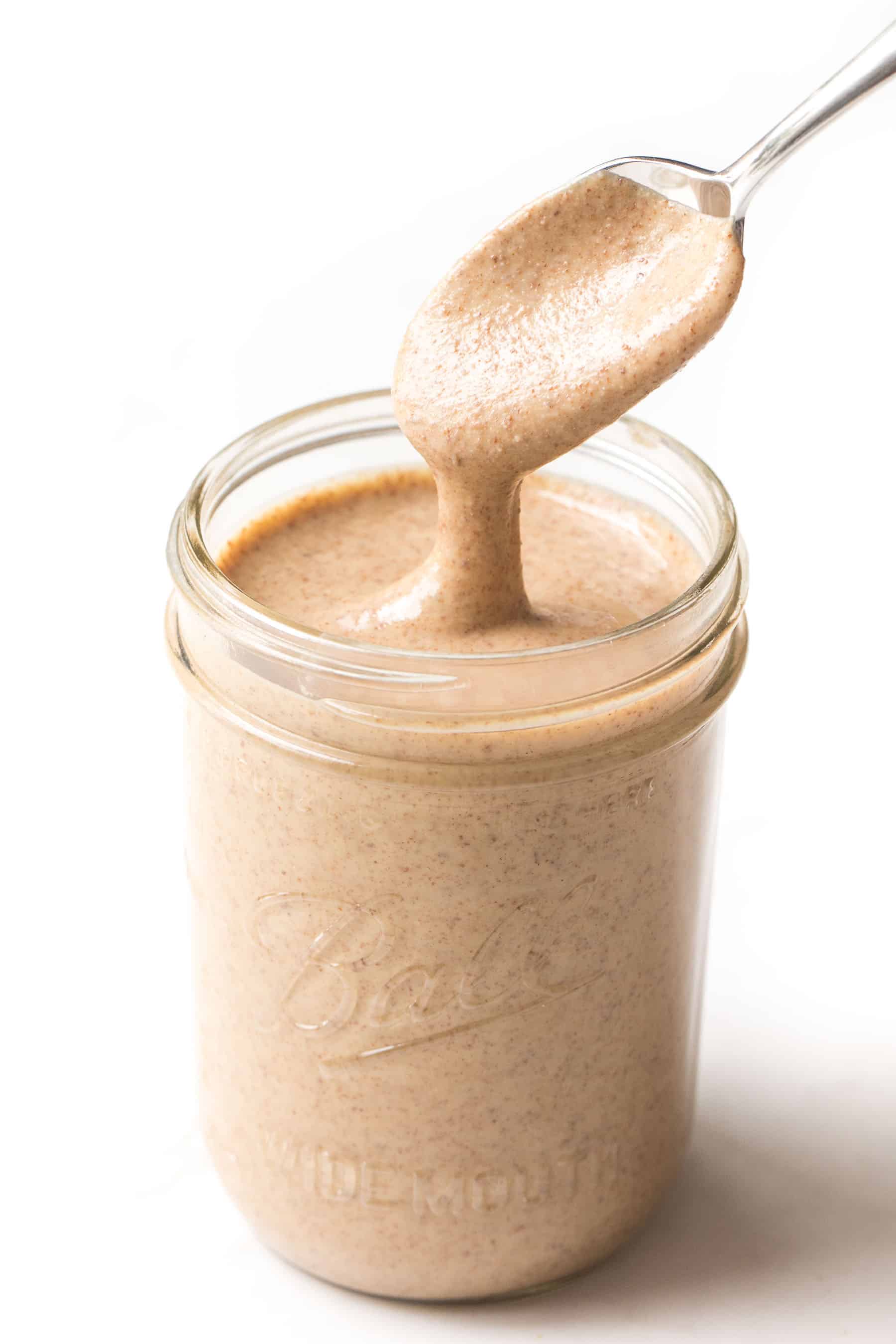 Spoon pouring homemade almond butter into a mason jar on a white background