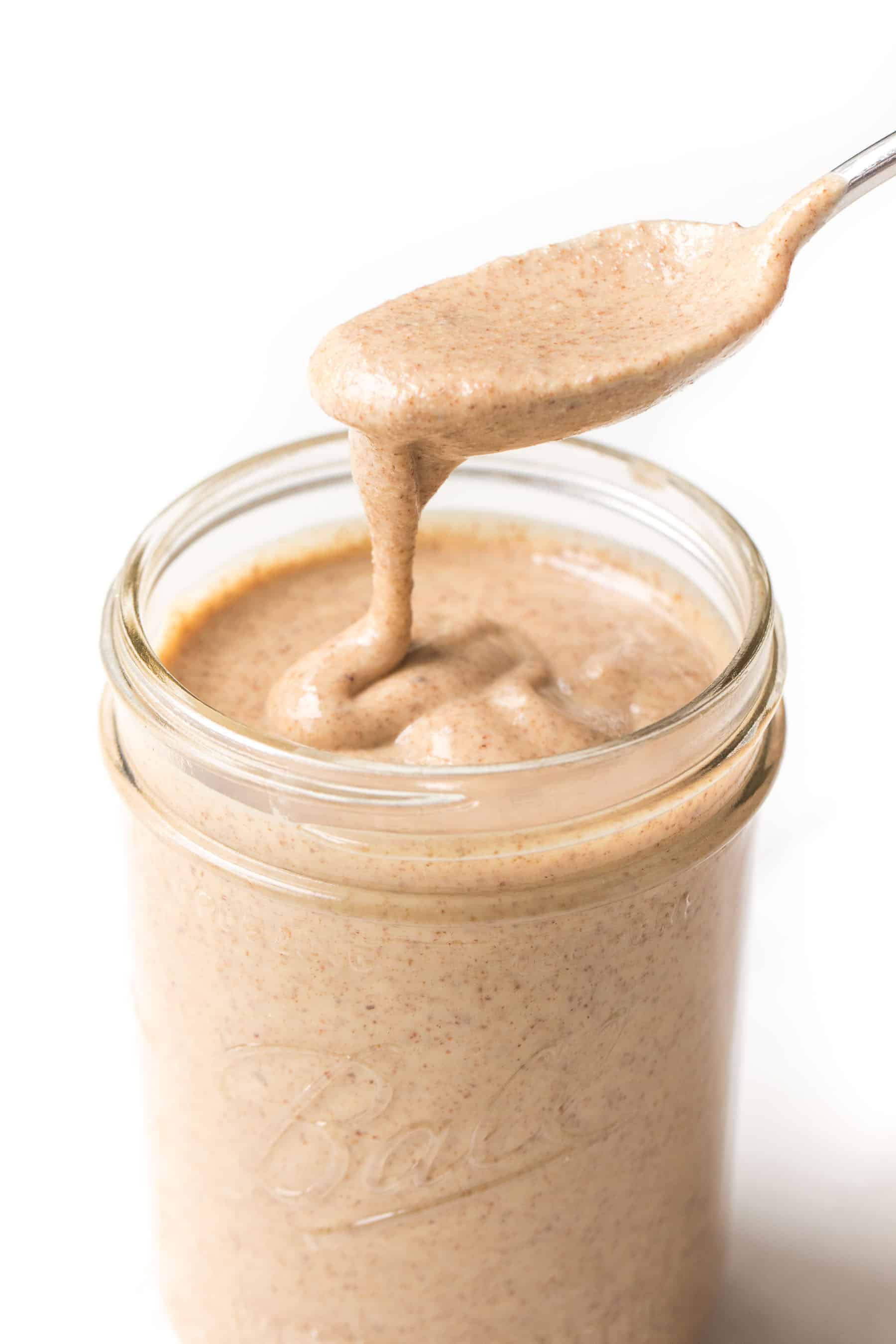 Spoon pouring homemade almond butter into a mason jar on a white background