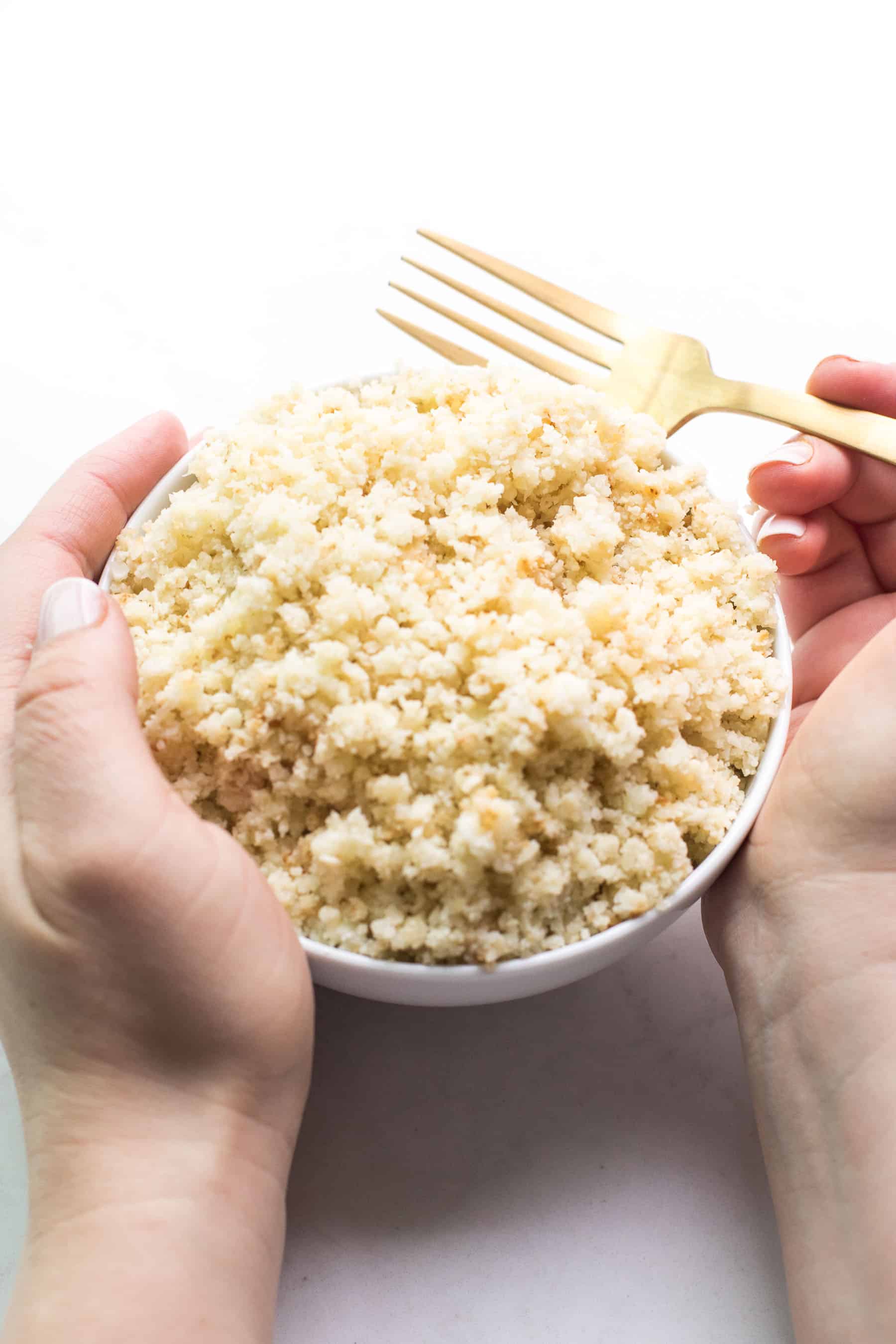 2 hands holding cauliflower rice in a white bowl with a fork