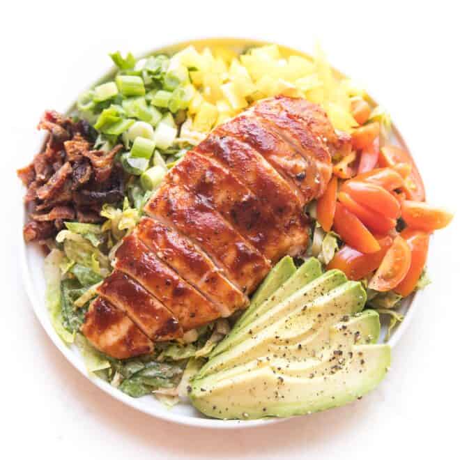 bbq chicken salad with avocado, bacon, bell pepper, tomatoes and green onions on a white plate and background
