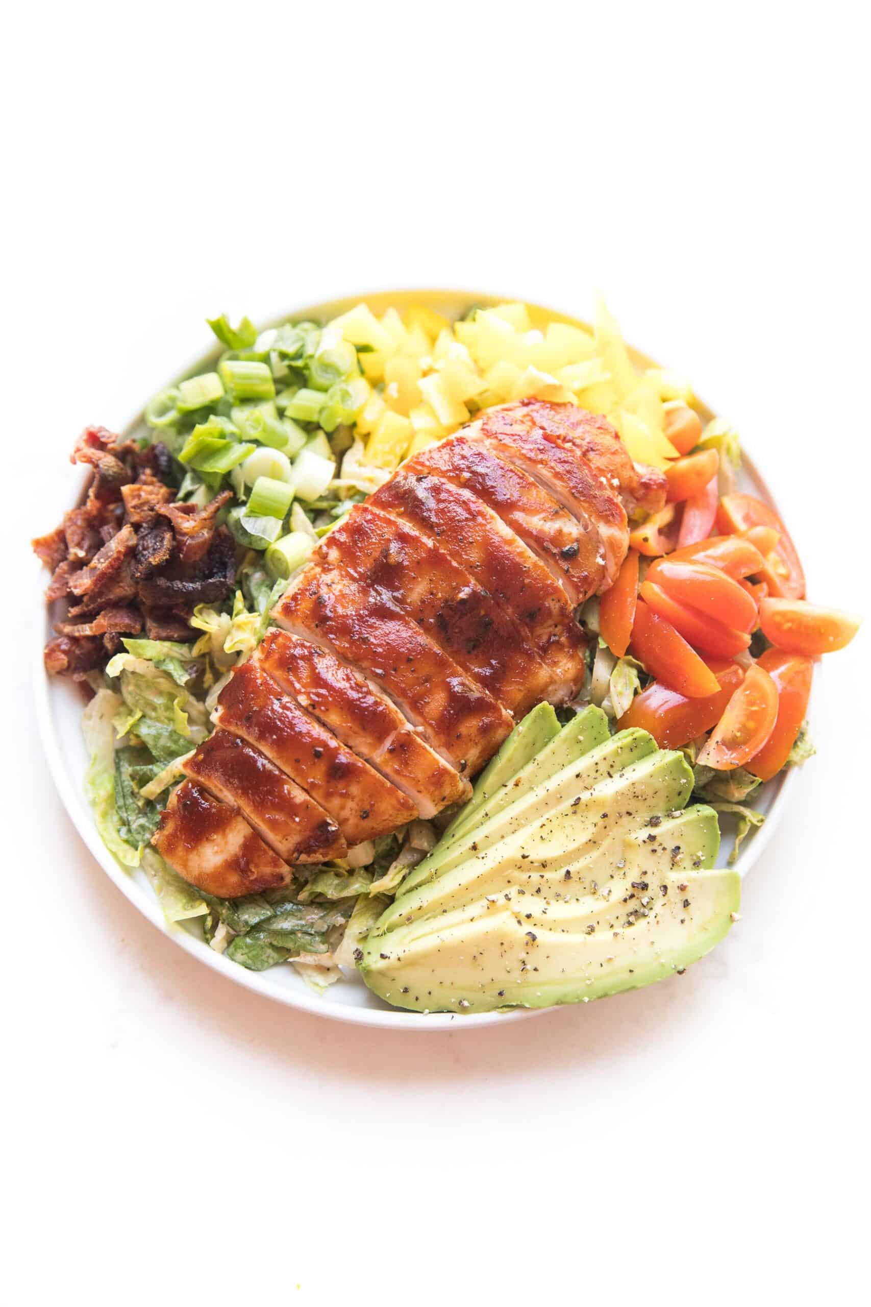 bbq chicken salad with avocado, bacon, bell pepper, tomatoes and green onions on a white plate and background