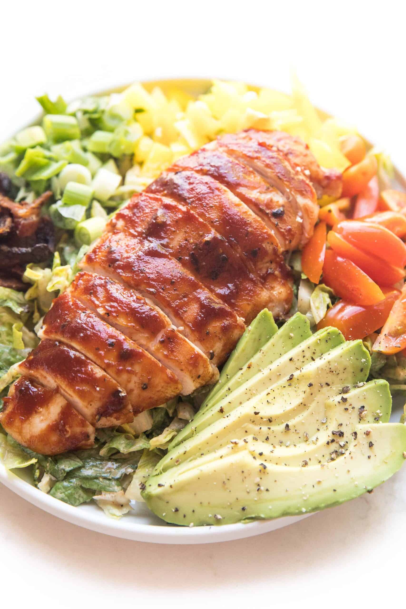 bbq chicken salad with avocado, bacon, bell pepper, tomatoes and green onions on a white plate and background