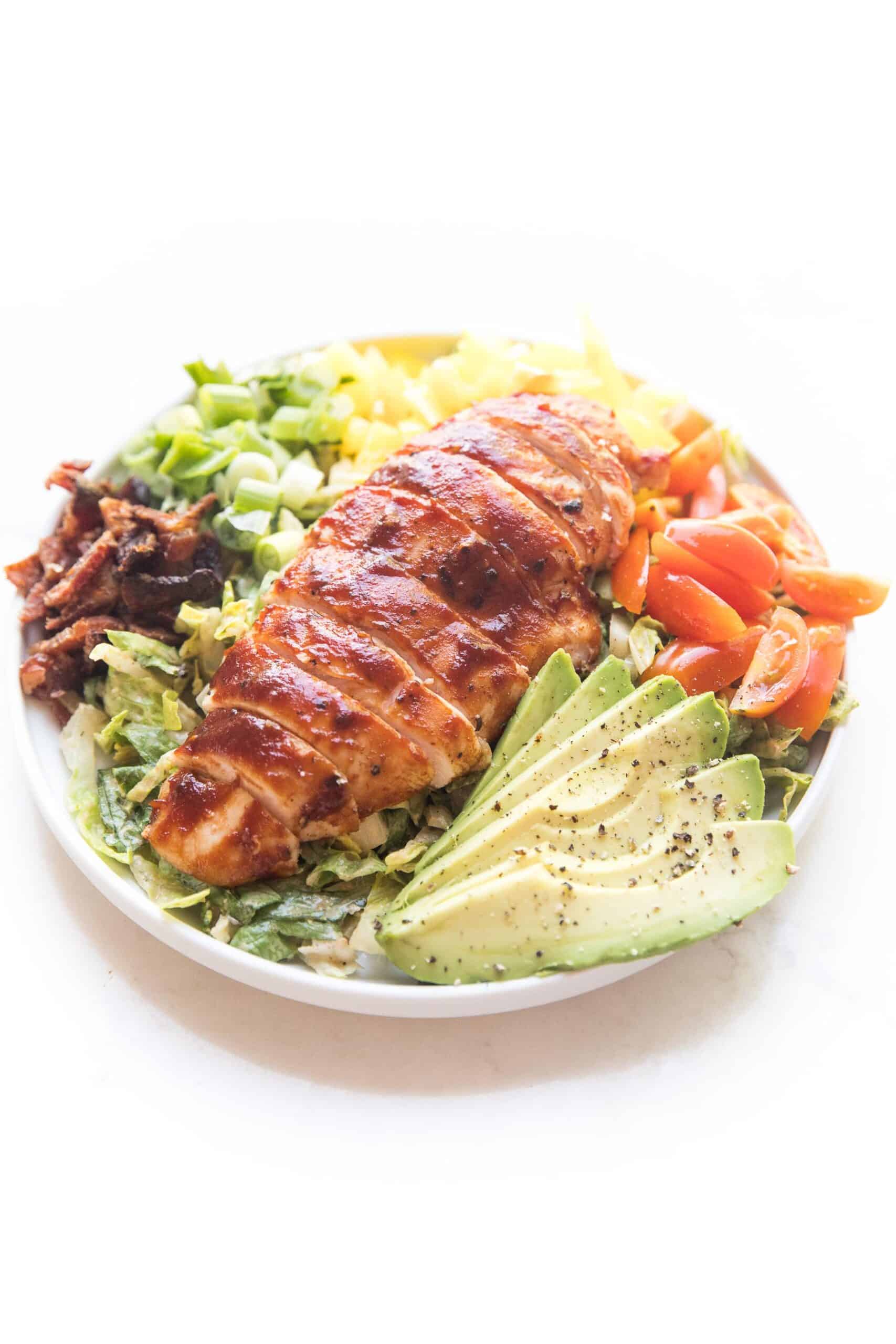 bbq chicken salad with avocado, bacon, bell pepper, tomatoes and green onions on a white plate and background