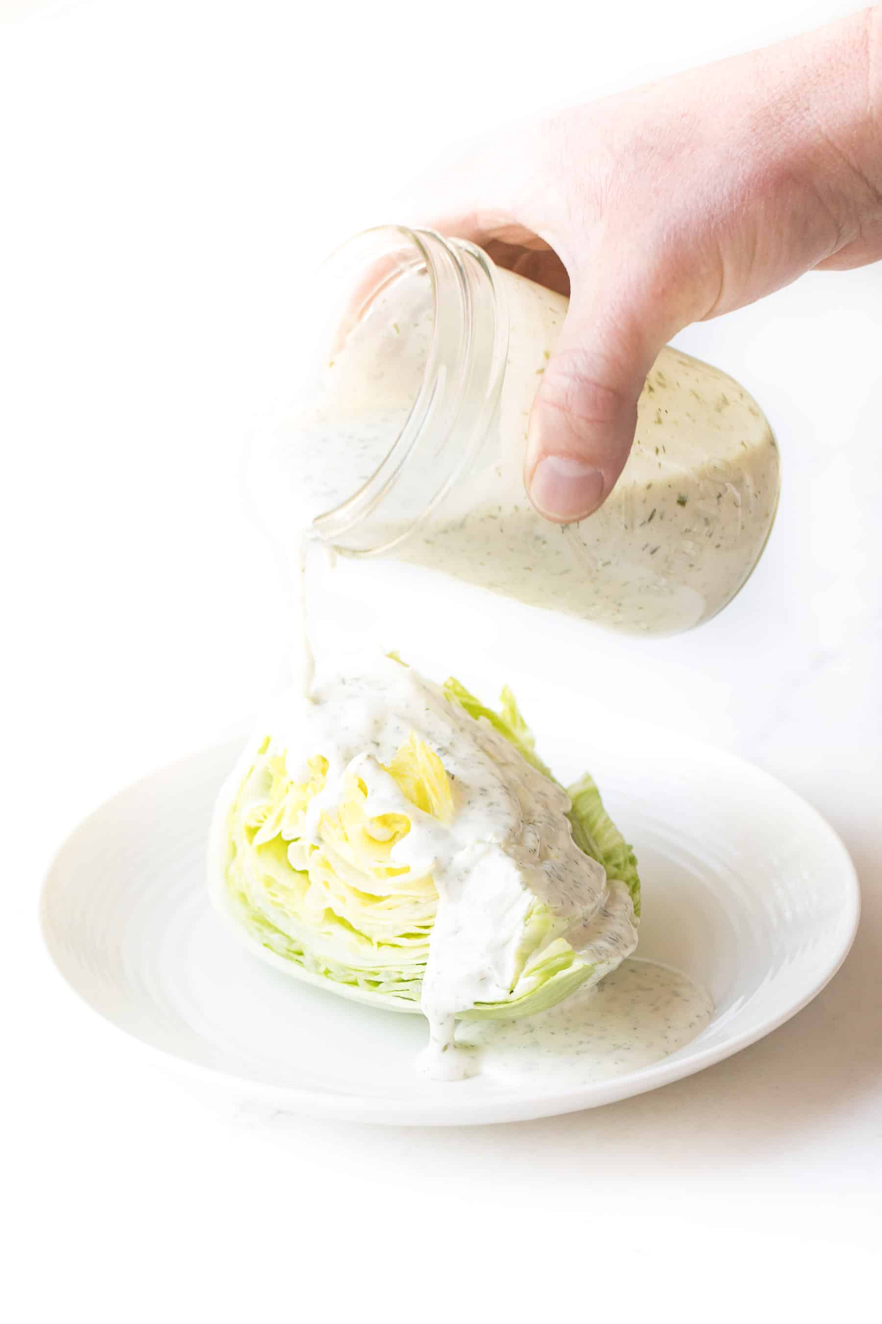 A jar of ranch being poured over iceberg lettuce on a white background