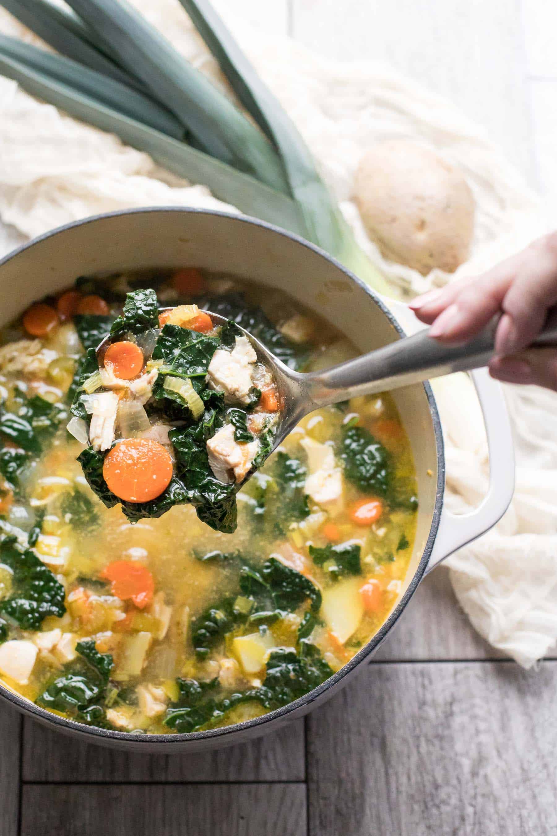 a large dutch oven filled with chicken kale soup with a hand pulling out a silver ladle of soup