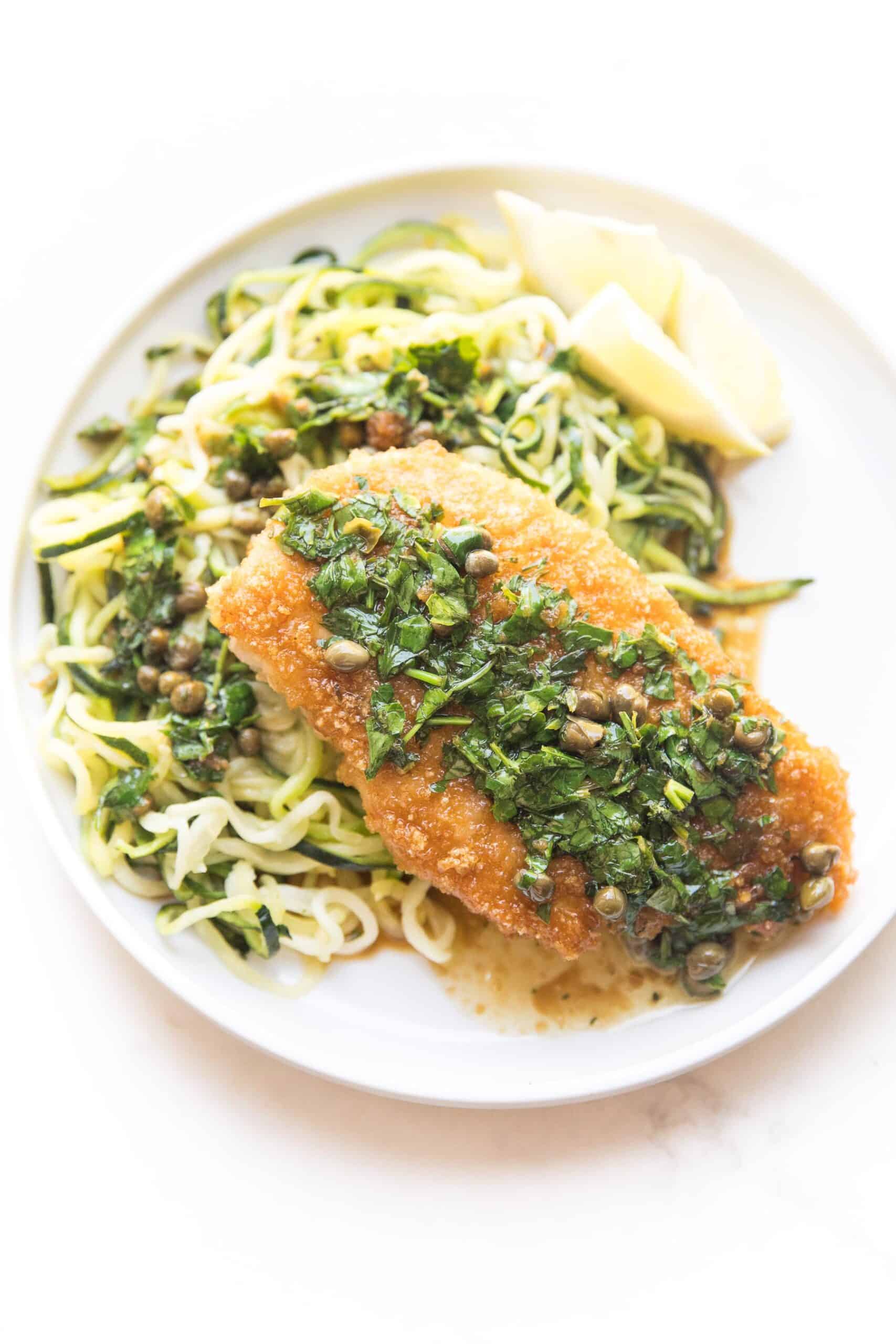 breaded lemon keto chicken piccata over zucchini noodles on a white plate and background