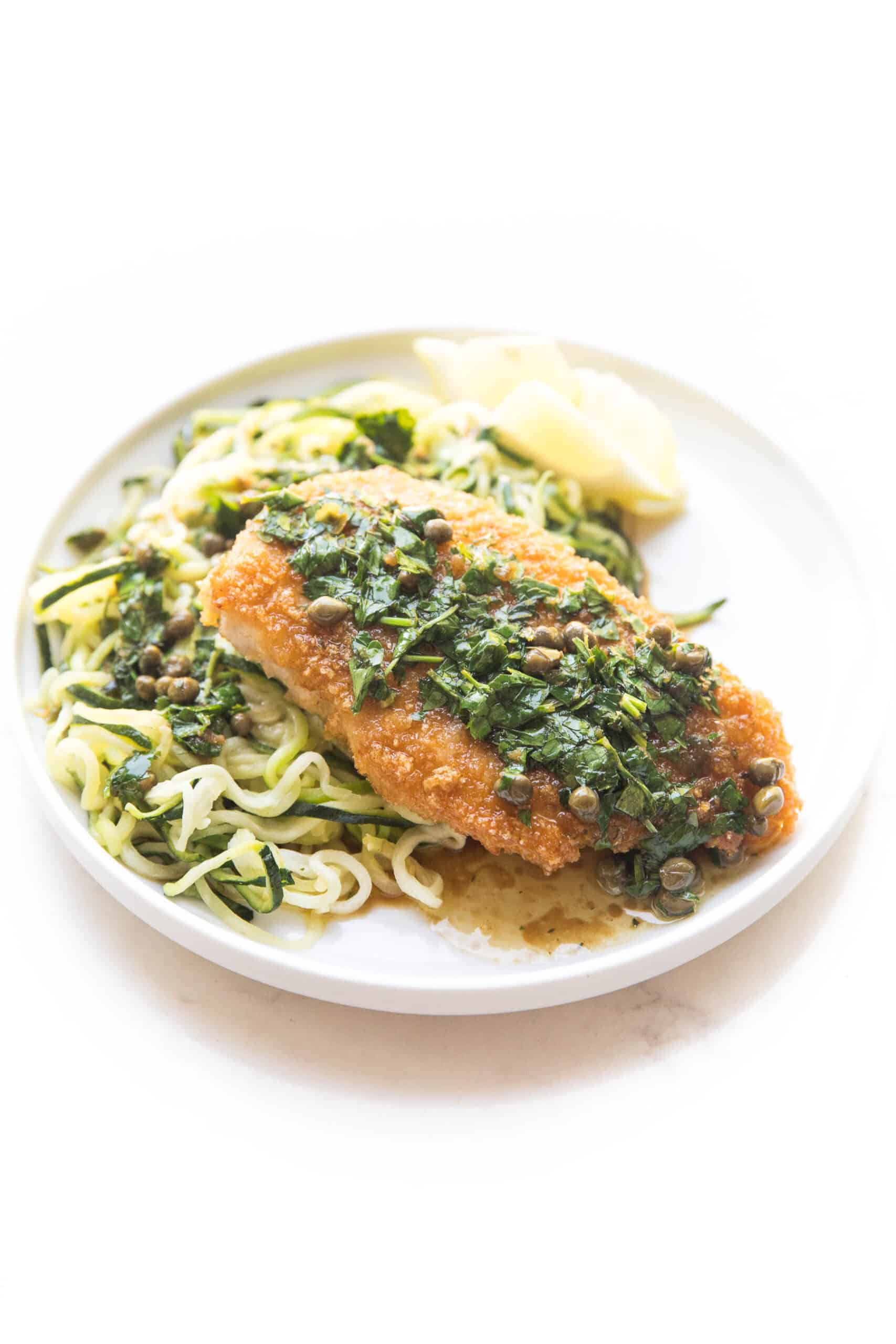 breaded lemon keto chicken piccata over zucchini noodles on a white plate and background