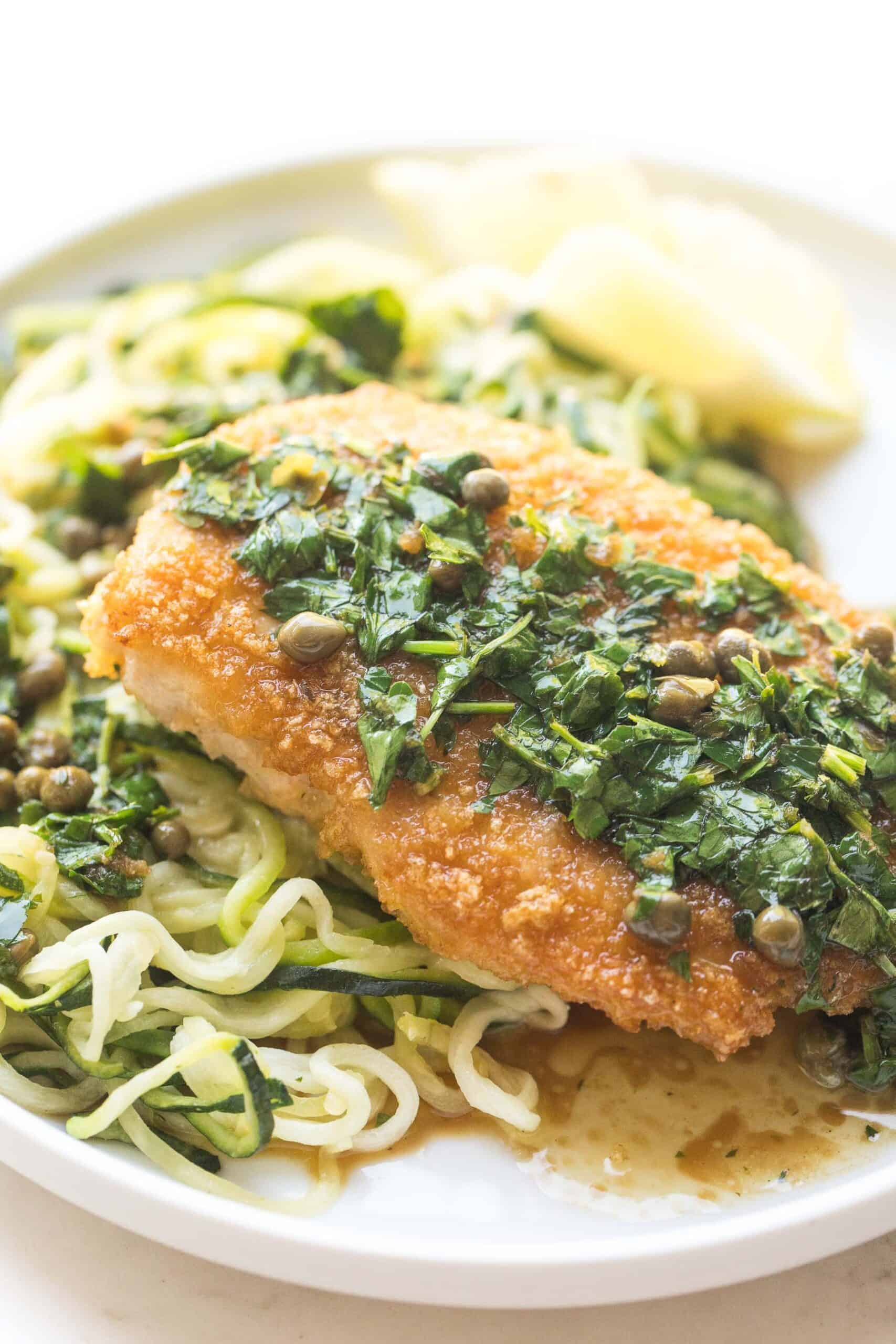 breaded lemon keto chicken piccata over zucchini noodles on a white plate and background