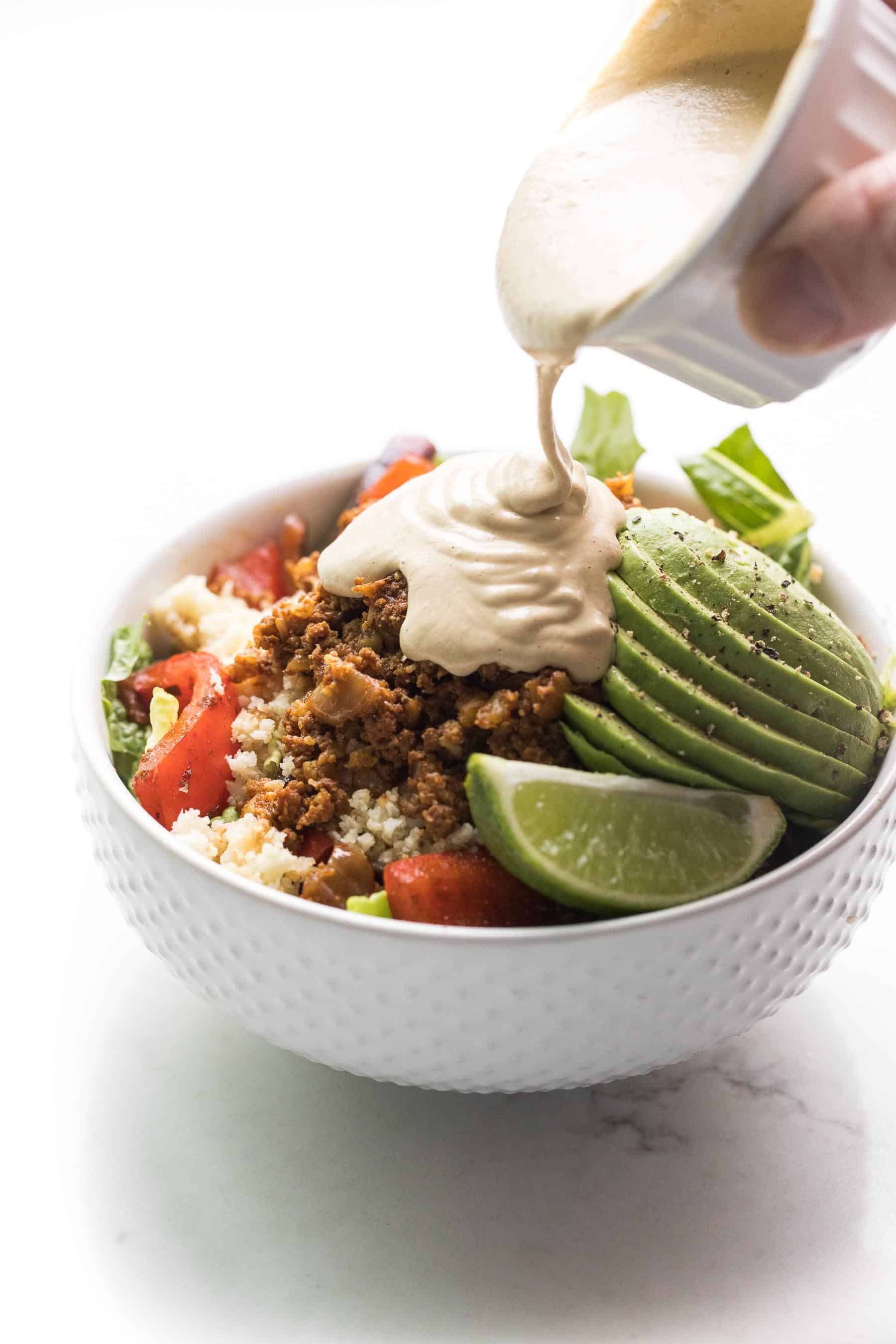 a hand pouring queso over a beef burrito bowl in a white bowl