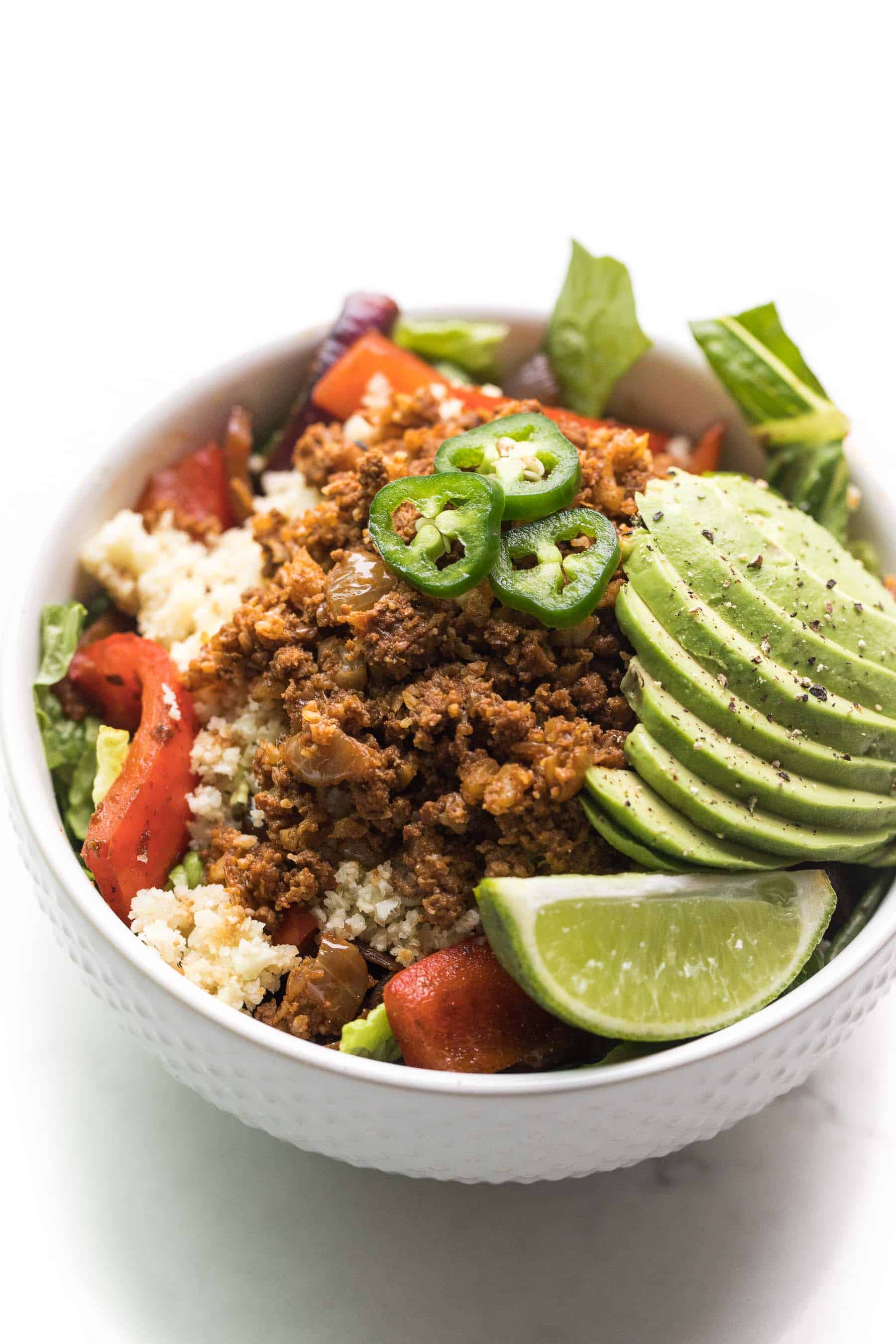 white bowl filled with ground beef, cauliflower rice, veggies, and lettuce, and topped with jalapeno, avocado, and lime
