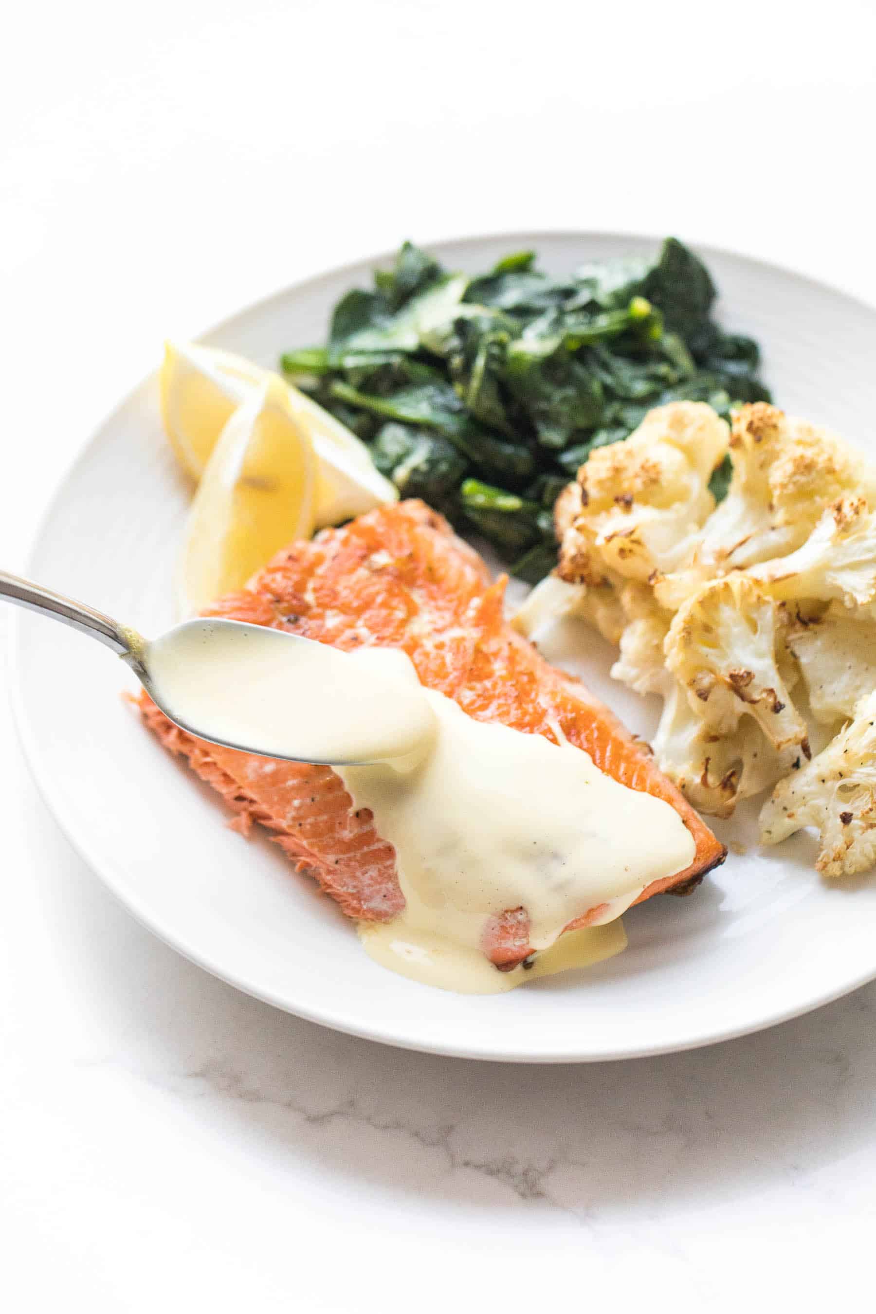 White plate with salmon and a spoon pouring hollandaise sauce, roasted cauliflower, sauteed spinach on a white background