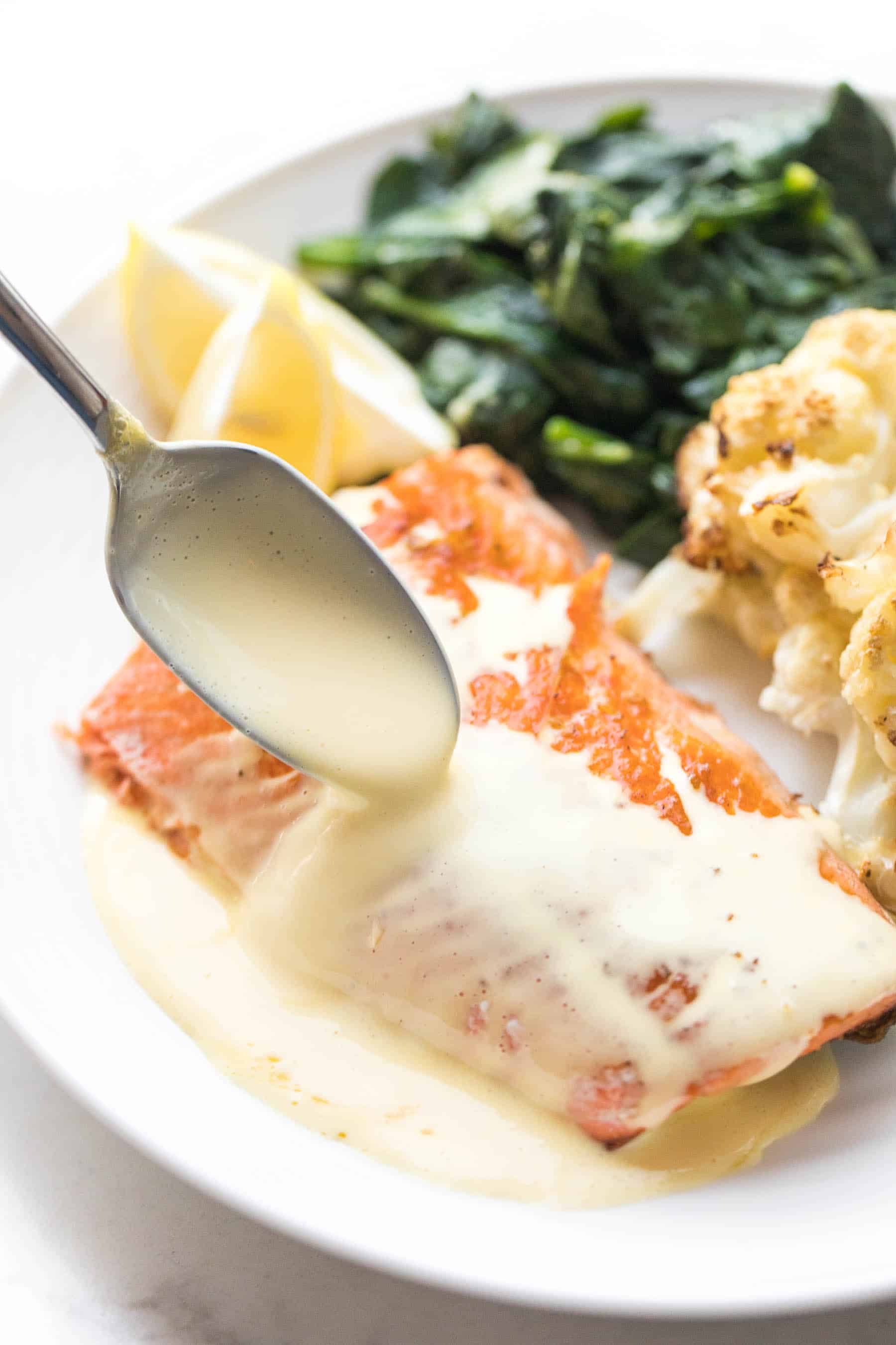 White plate with salmon and a spoon pouring hollandaise sauce, roasted cauliflower, sauteed spinach on a white background