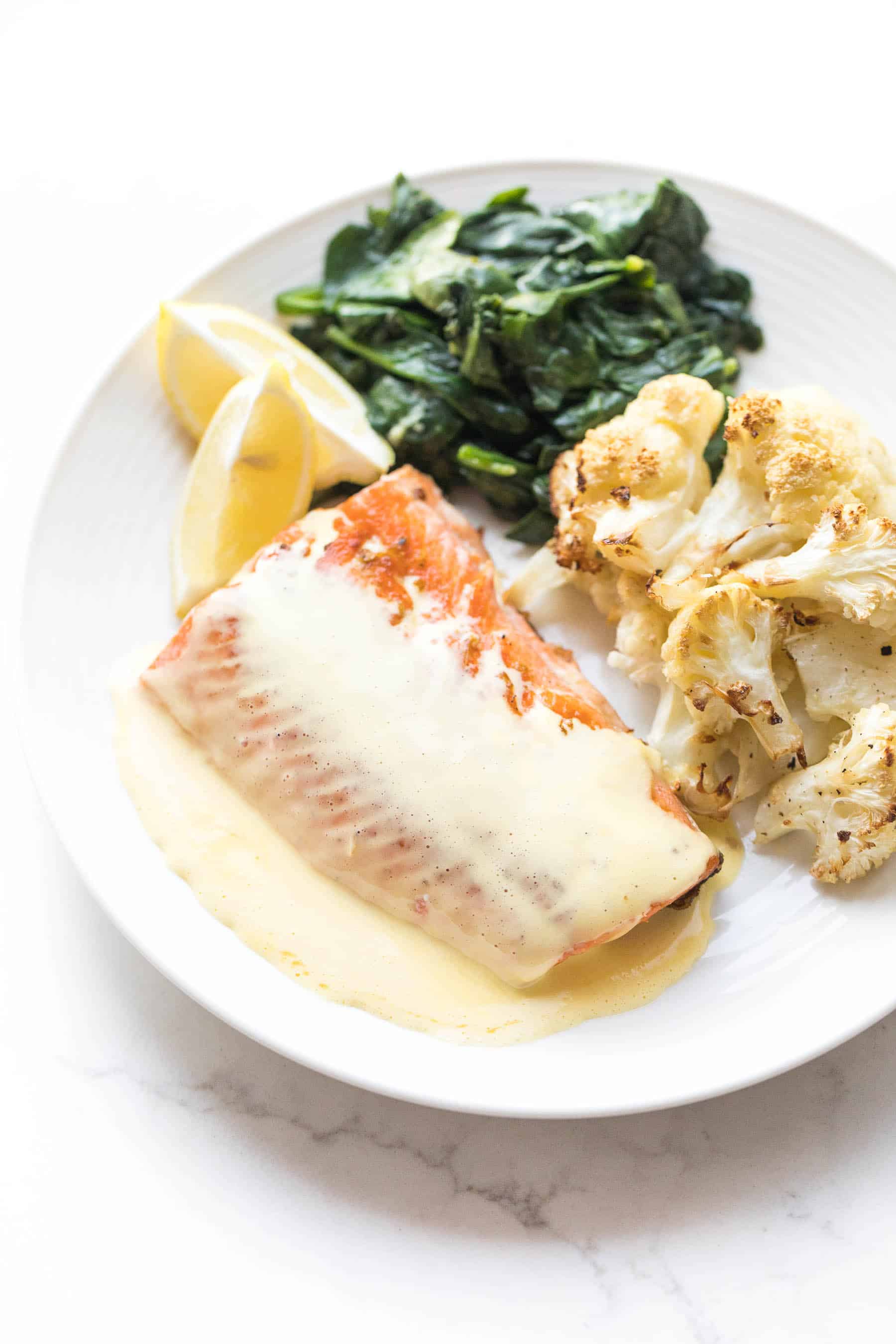 White plate with salmon and hollandaise sauce, roasted cauliflower, sauteed spinach on a white background