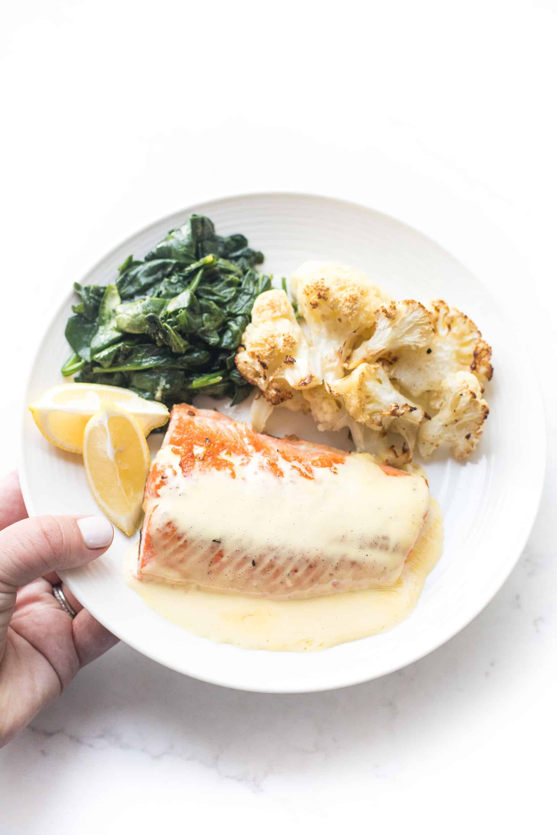 a hand holding a white plate with salmon and hollandaise sauce, roasted cauliflower, sauteed spinach on a white background