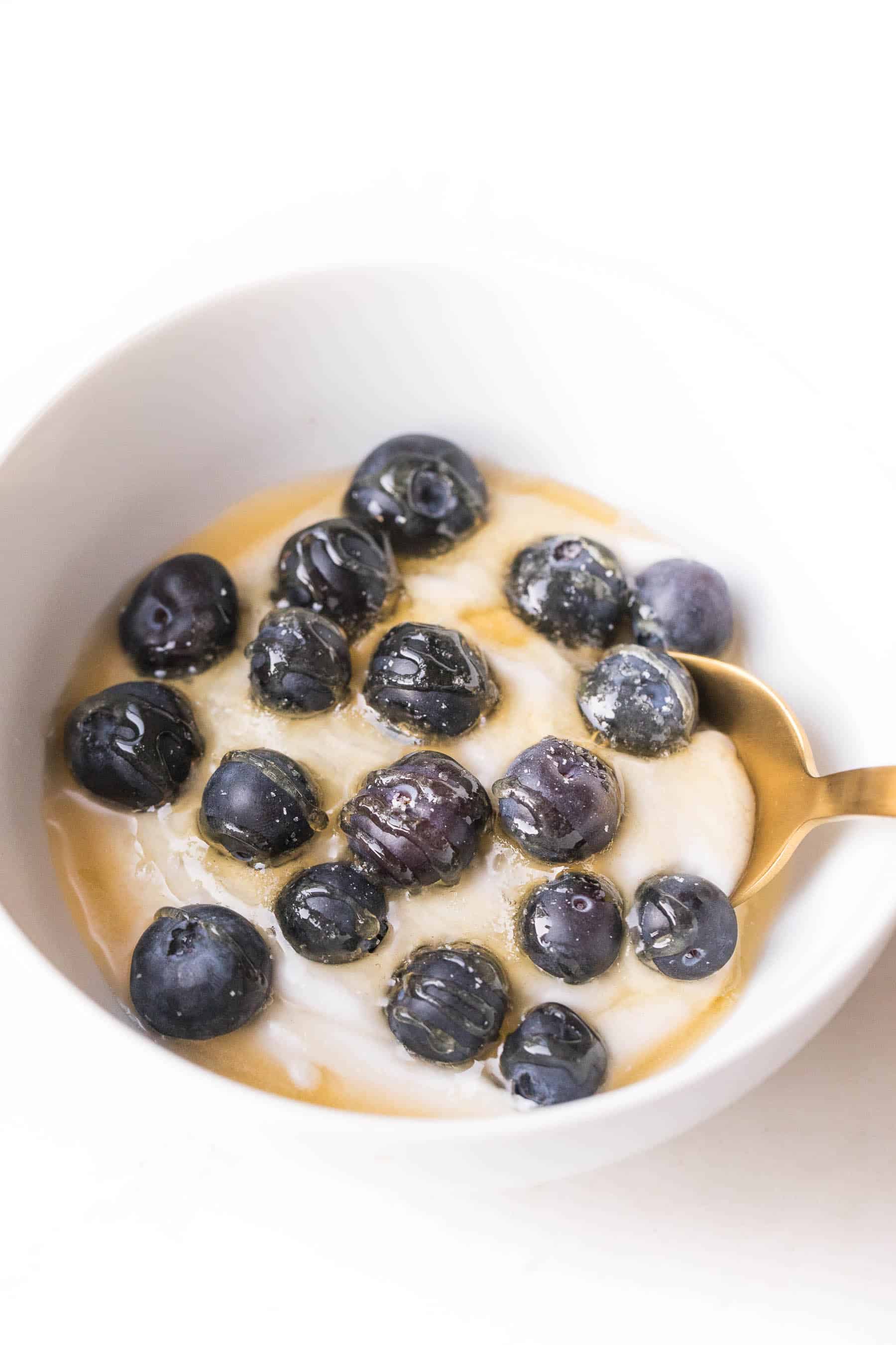 white bowl on a white background with coconut whipped cream, blueberries and honey