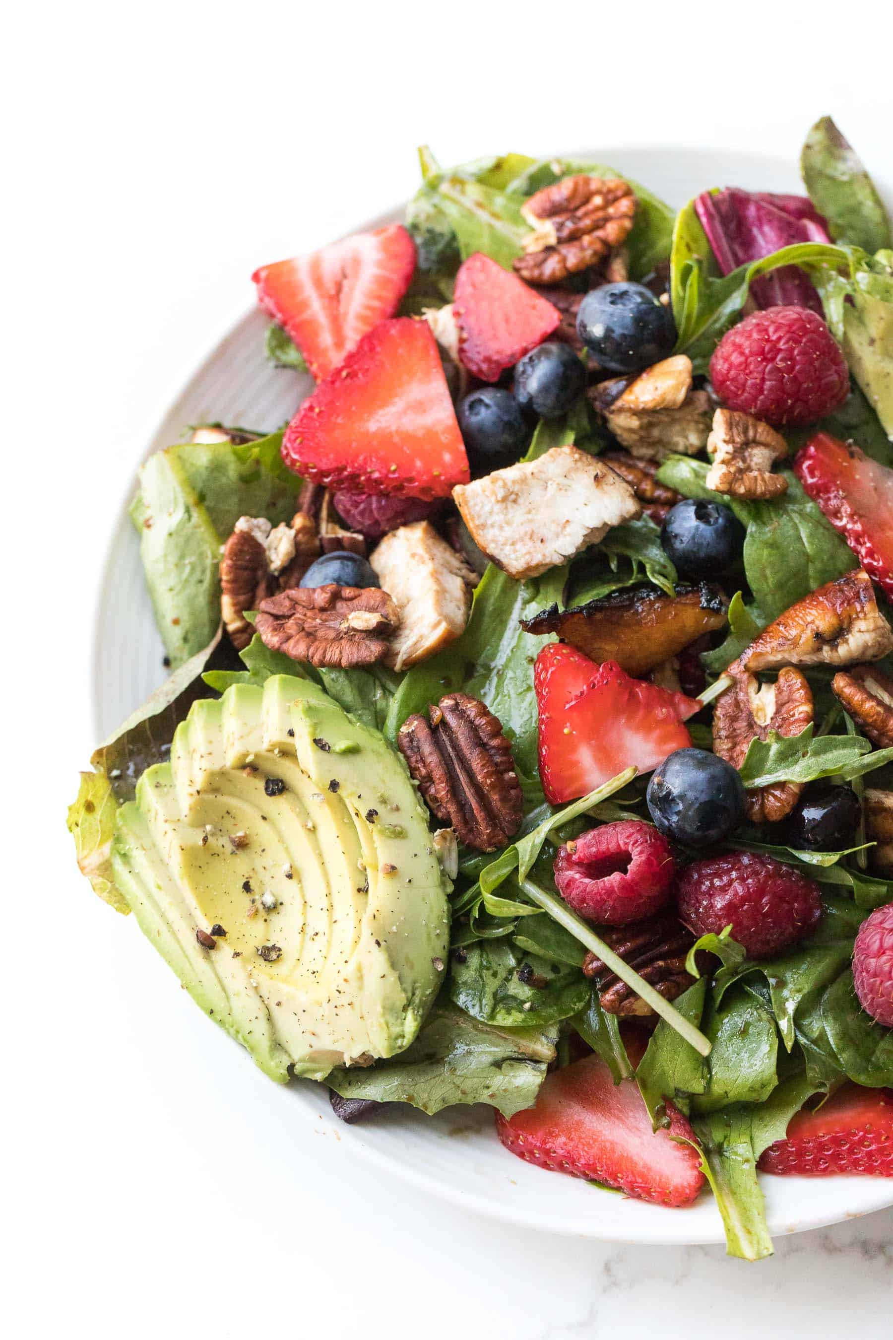 salad with strawberries, blueberries, raspberries, chicken, avocado + pecans on a white plate and background