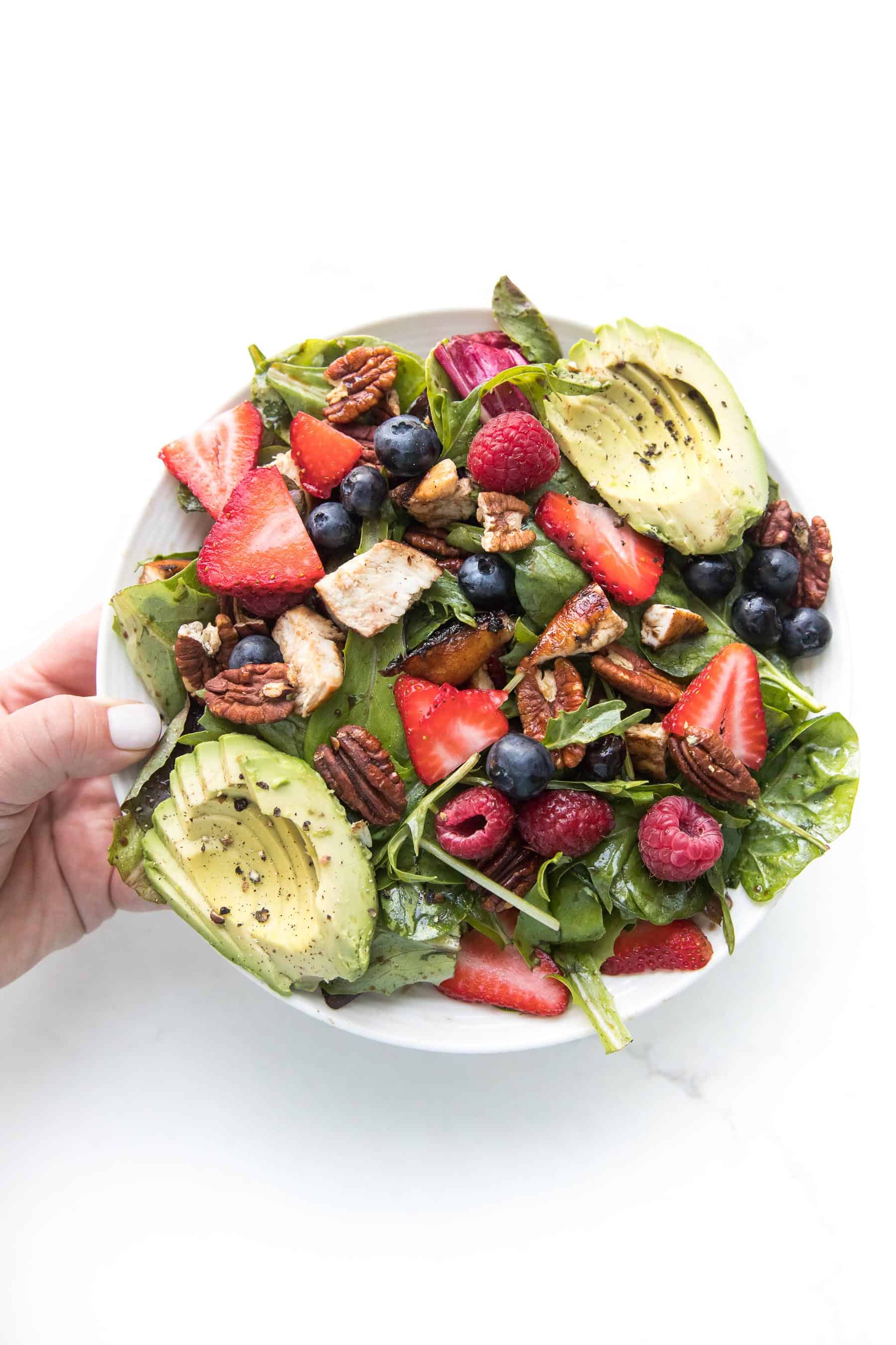 hand holding a salad with strawberries, blueberries, raspberries, chicken, avocado + pecans on a white plate and background