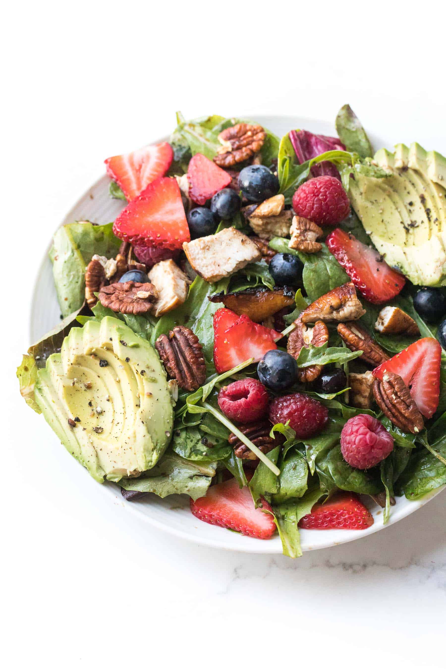 salad with strawberries, blueberries, raspberries, chicken, avocado + pecans on a white plate and background