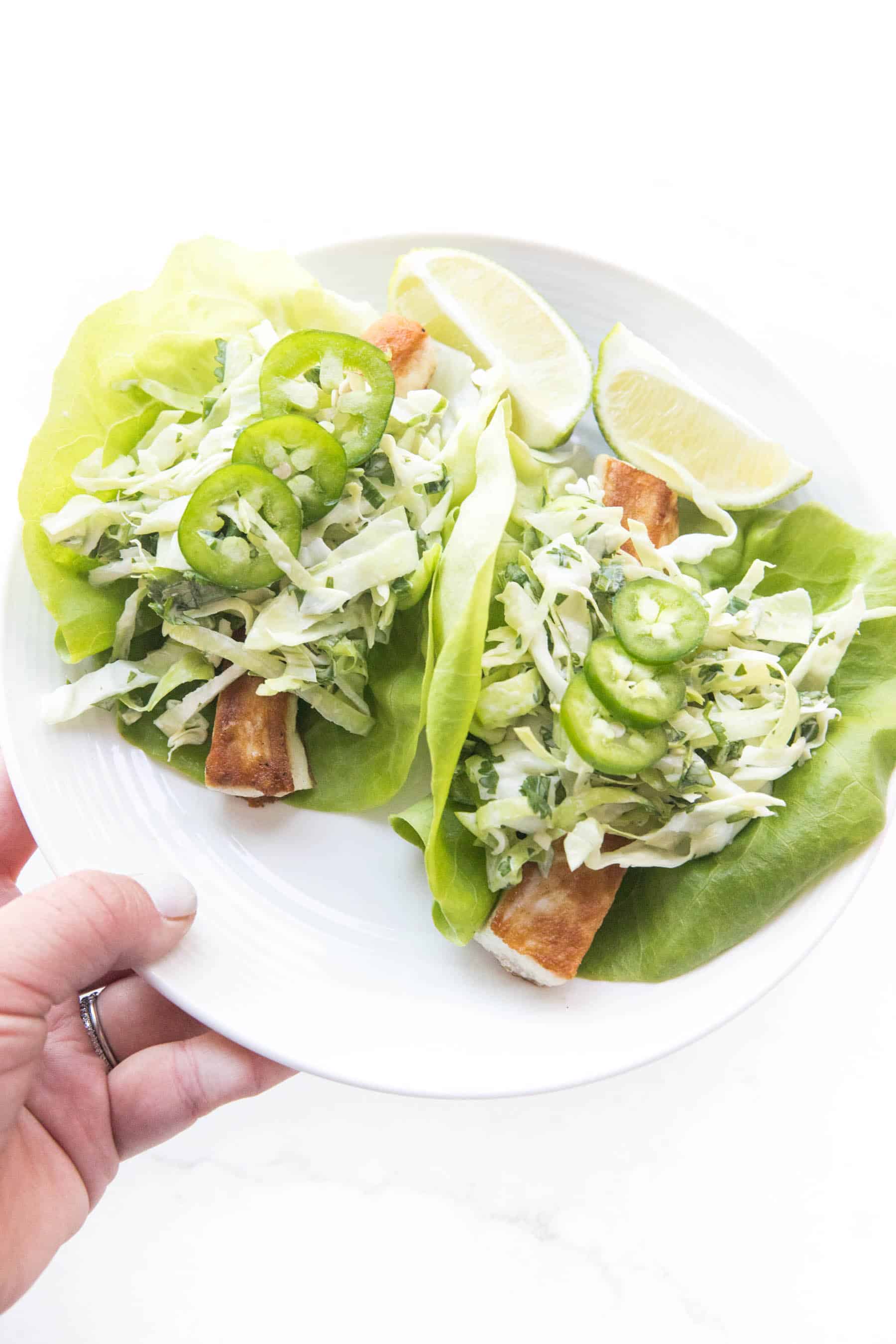 hand holding fish tacos on a lettuce taco shell topped with coleslaw on a white background