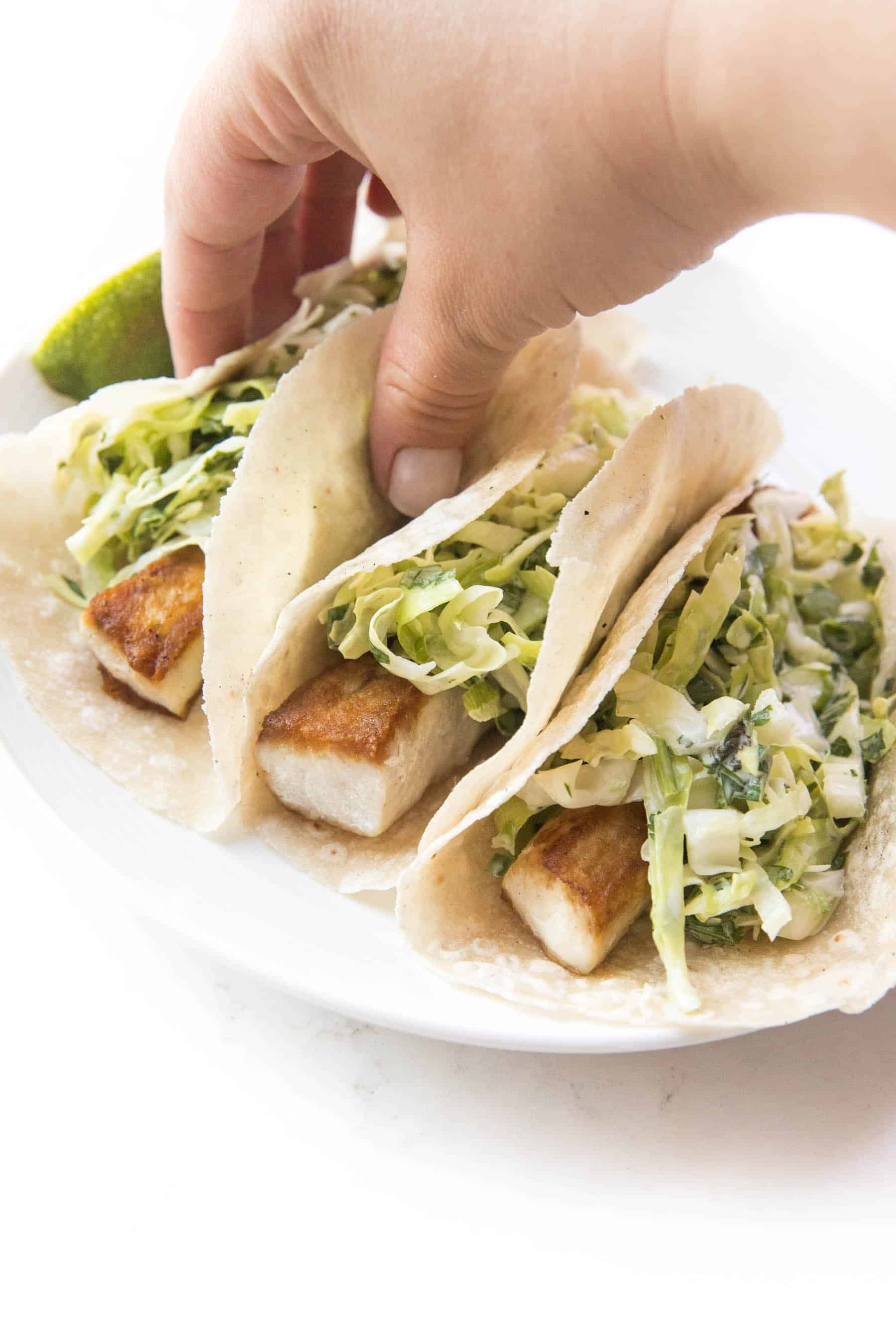 hand grabbing a fish tacos topped with coleslaw on a white background