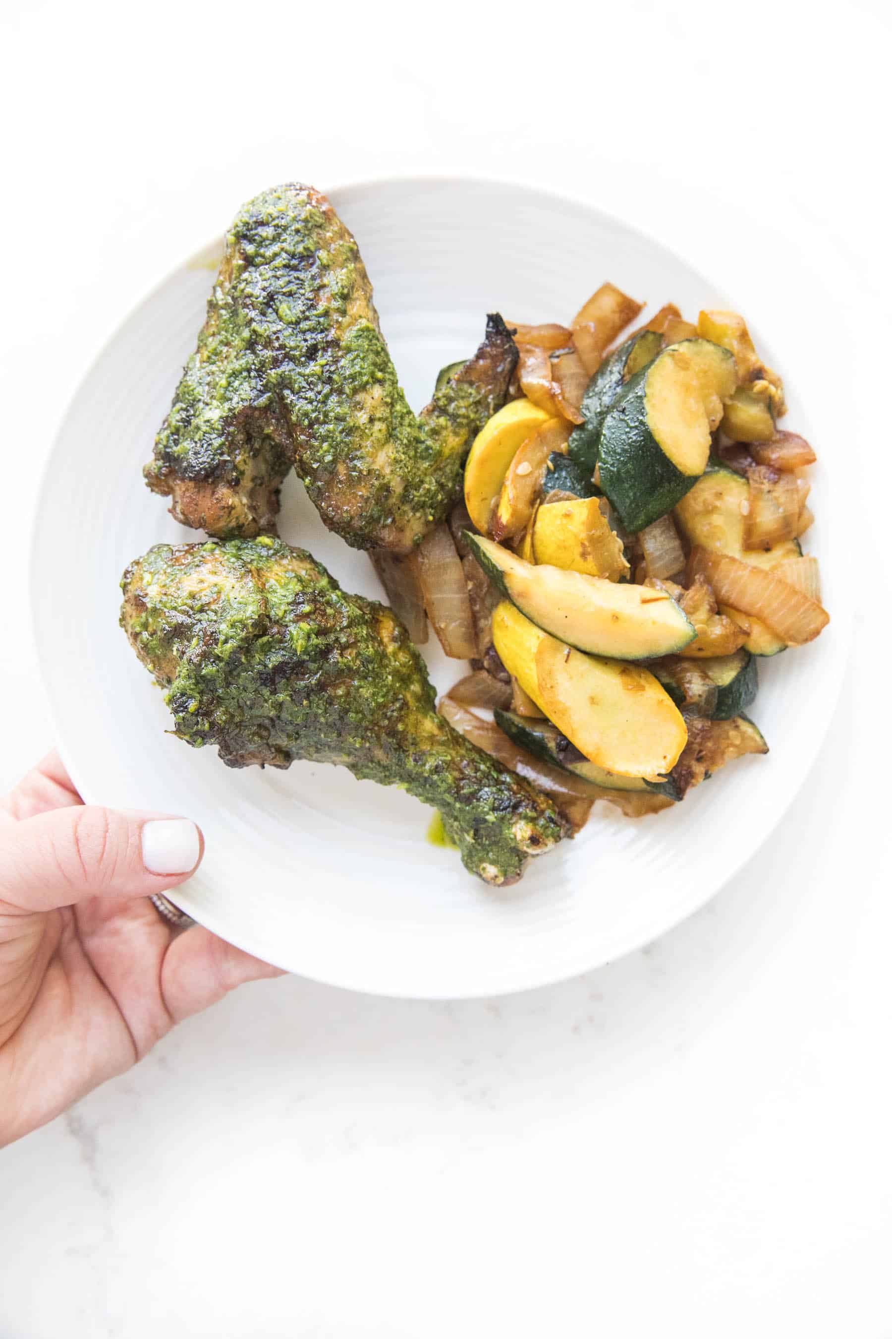 hand holding a white plate of grilled chicken with chimichurri chicken and squash on a white background