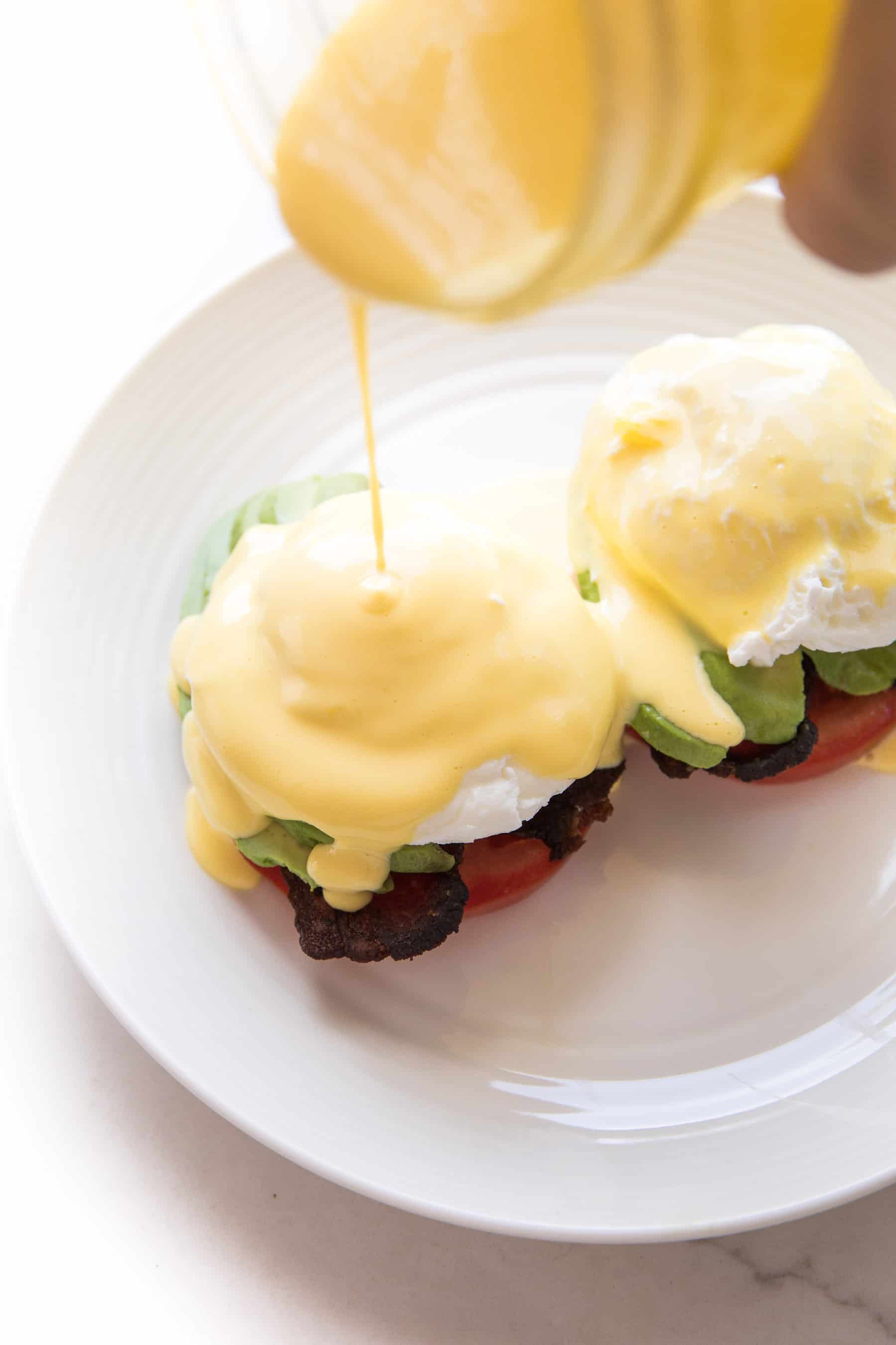 hollandaise sauce being poured over eggs benedict on a white plate