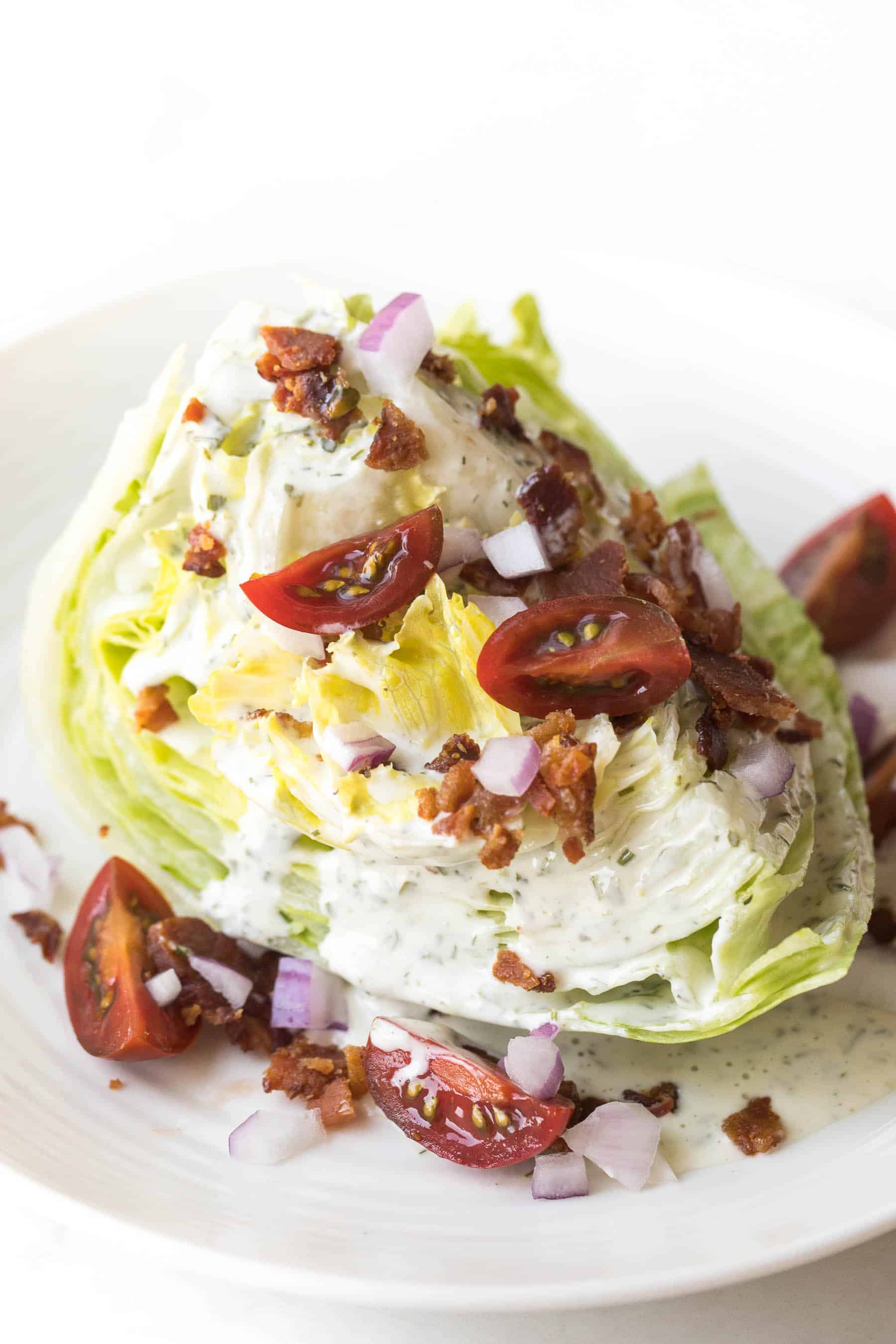 wedge salad topped with dressing, bacon, tomatoes and onion on a white plate and white background