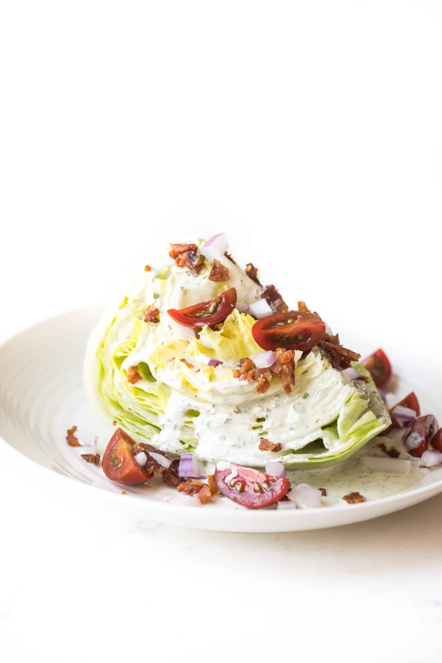 wedge salad topped with dressing, bacon, tomatoes and onion on a white plate and white background