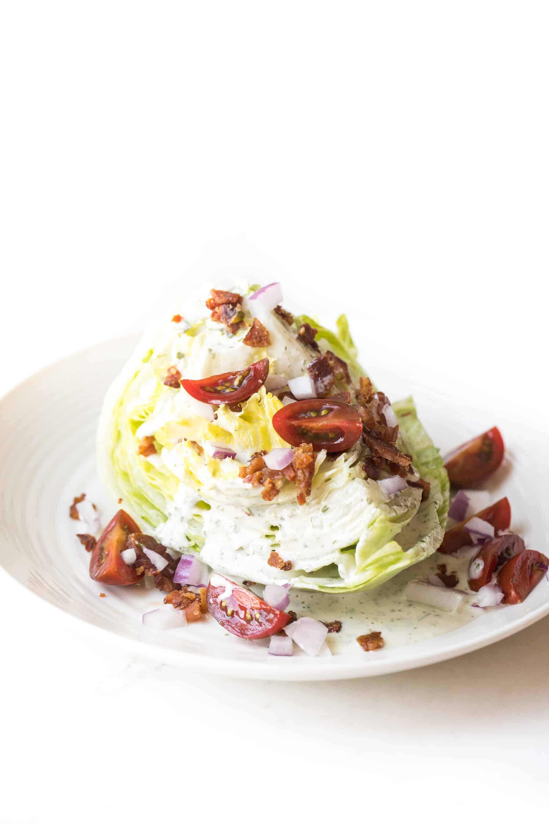 wedge salad topped with dressing, bacon, tomatoes and onion on a white plate and white background