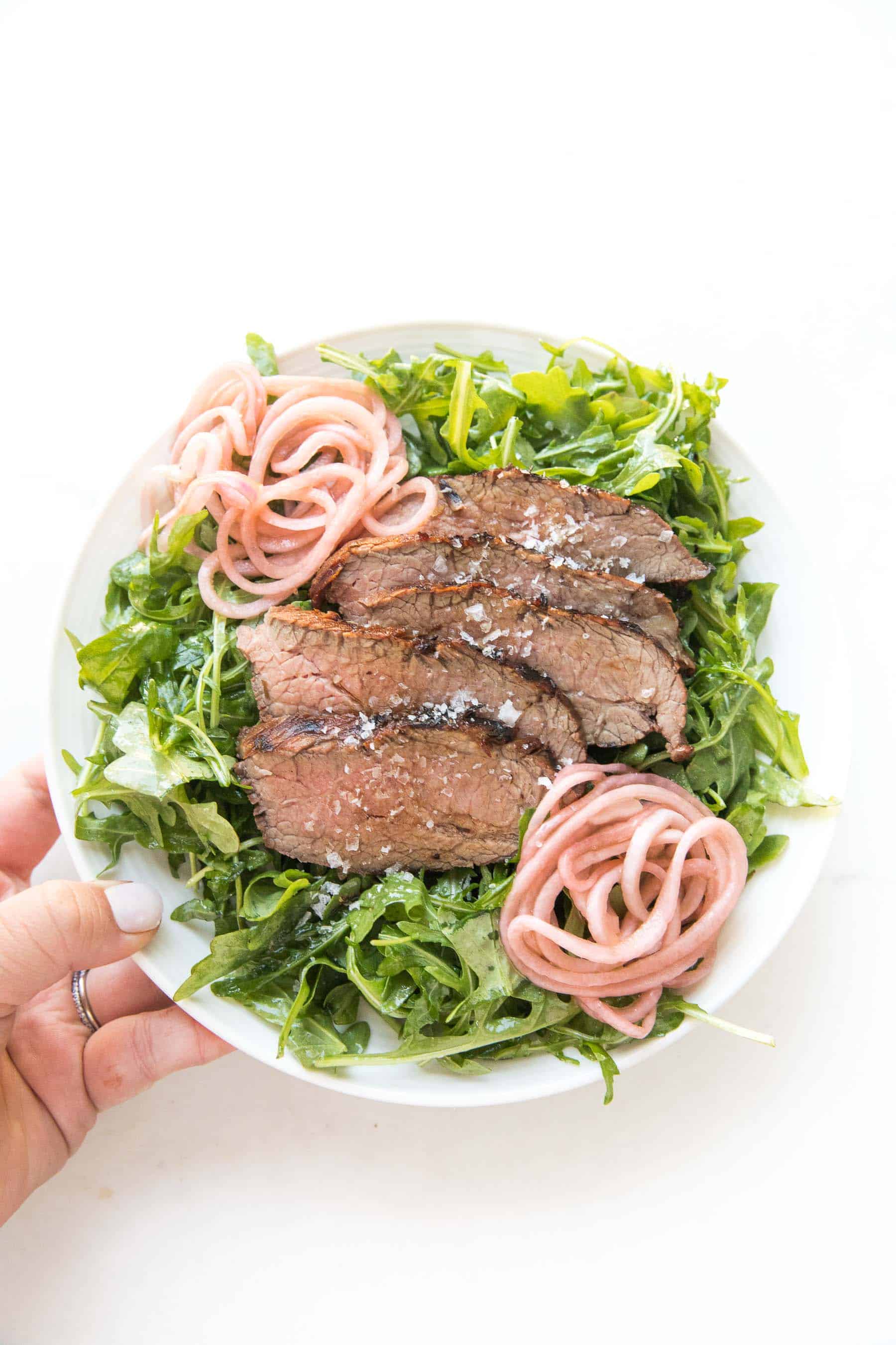 hand holding Tri tip arugula salad with pickled onions on a white plate and white background