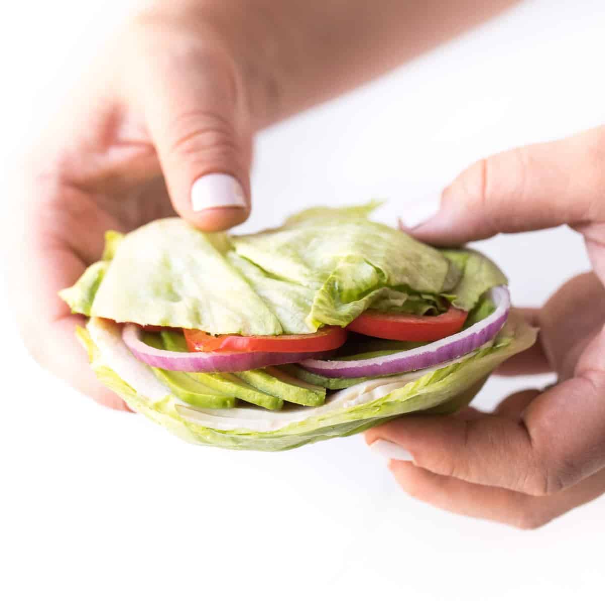 a hand holding two slices of iceberg lettuce as a "bun" with avocado, tomato, and red onions inside
