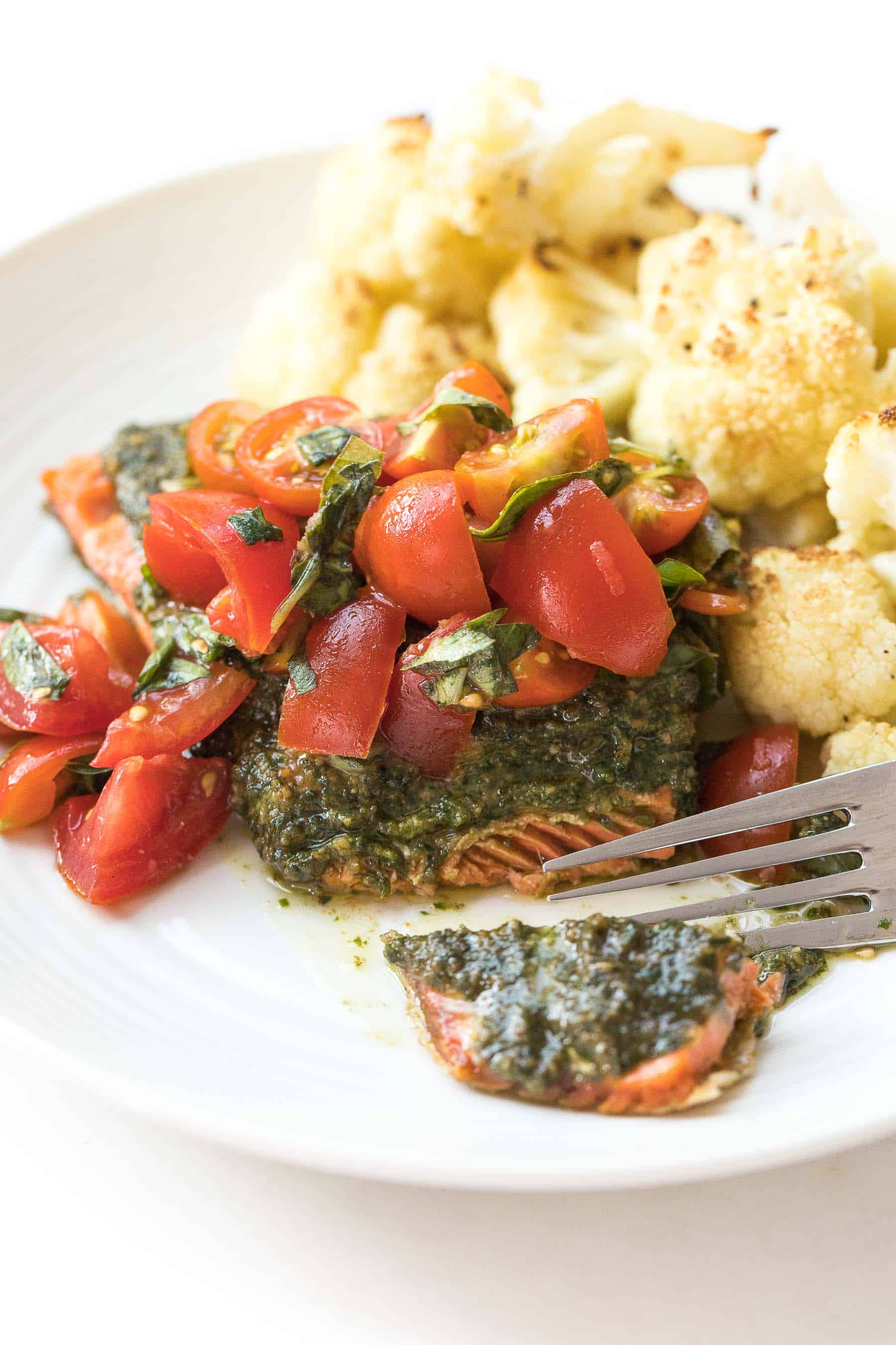 fork cutting salmon topped with pesto and tomatoes on a white plate and white background