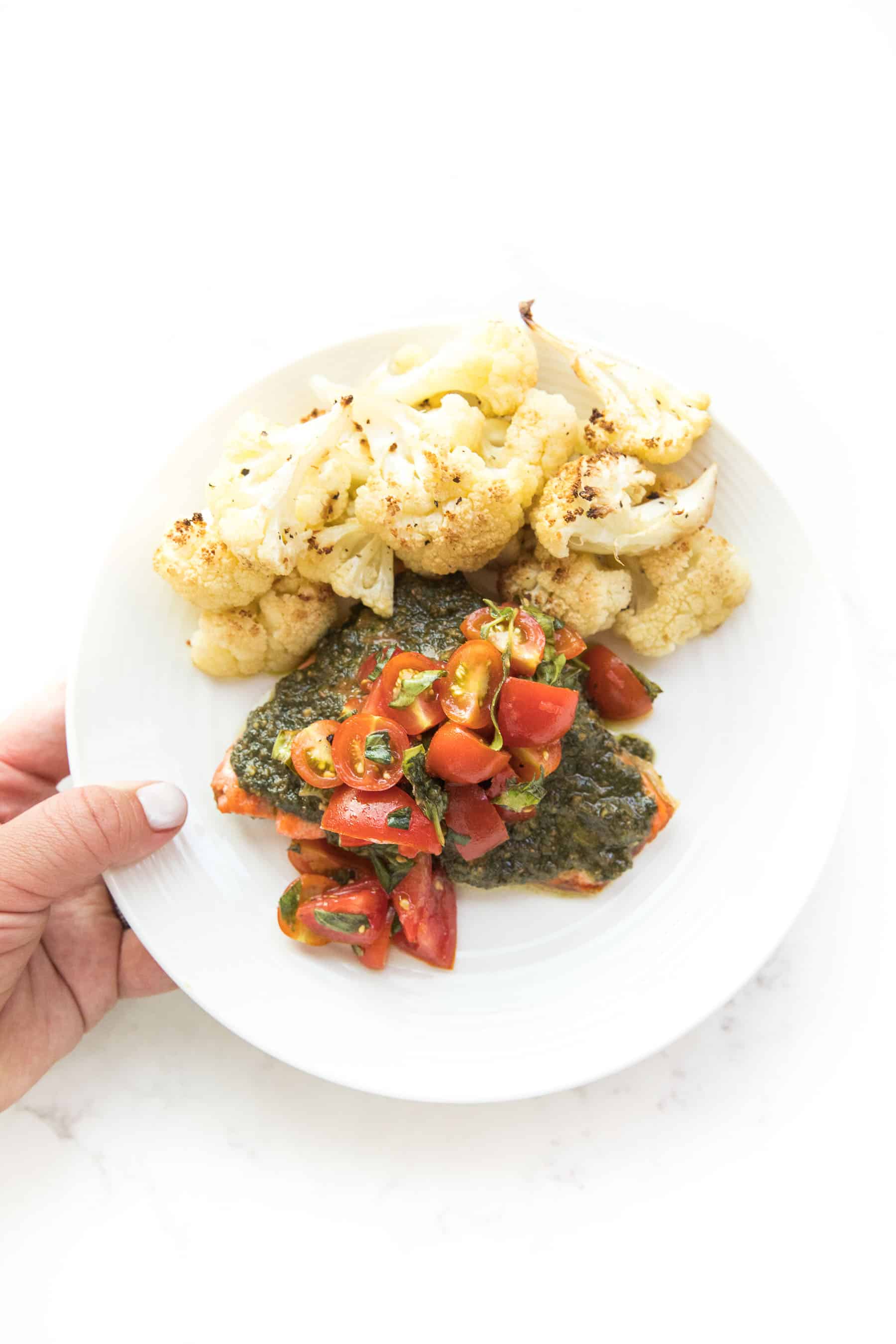 hand holding salmon topped with pesto and tomatoes on a white plate and white background