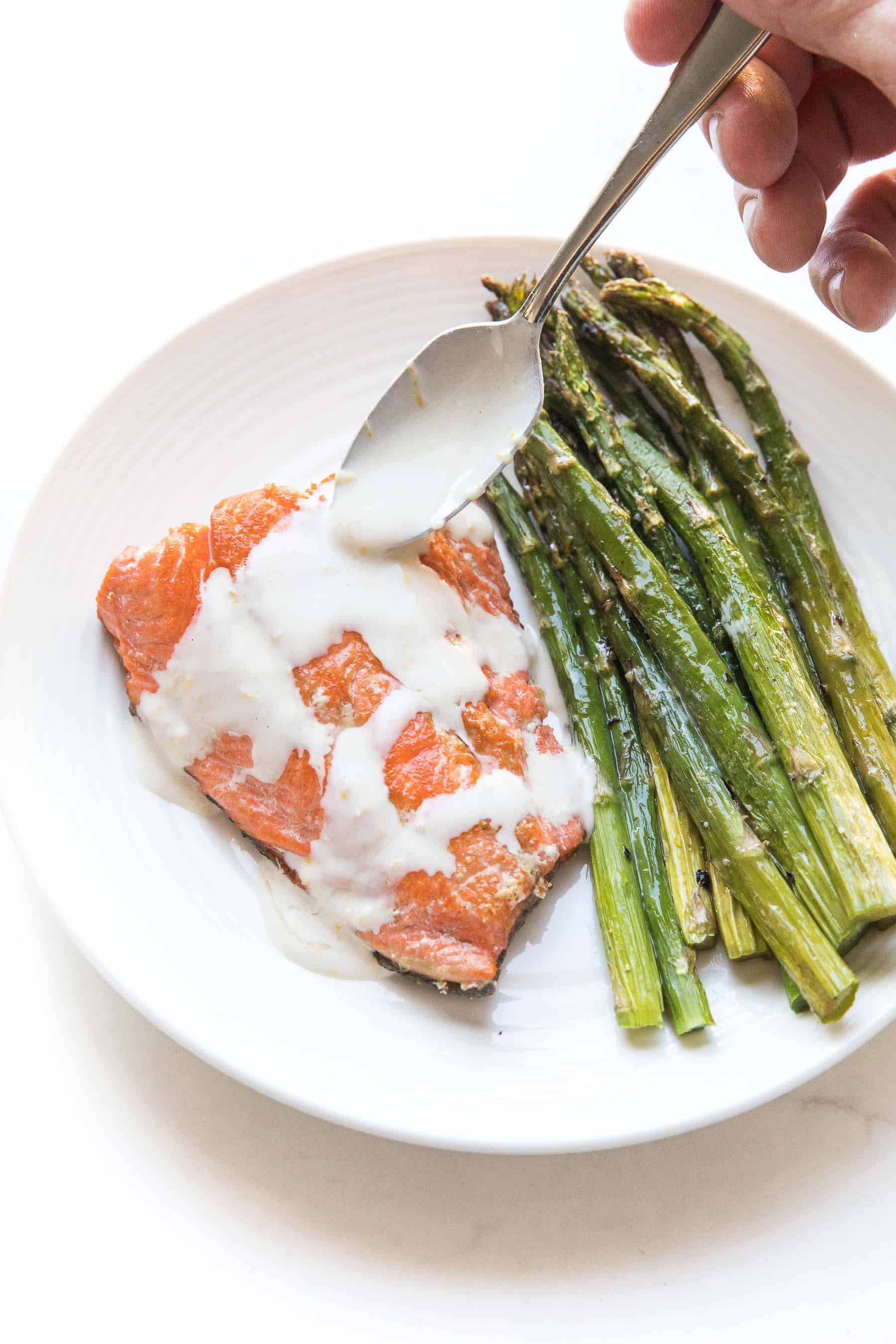 spoon drizzling Salmon + asparagus with a white creamy coconut tahini sauce on a white plate and background