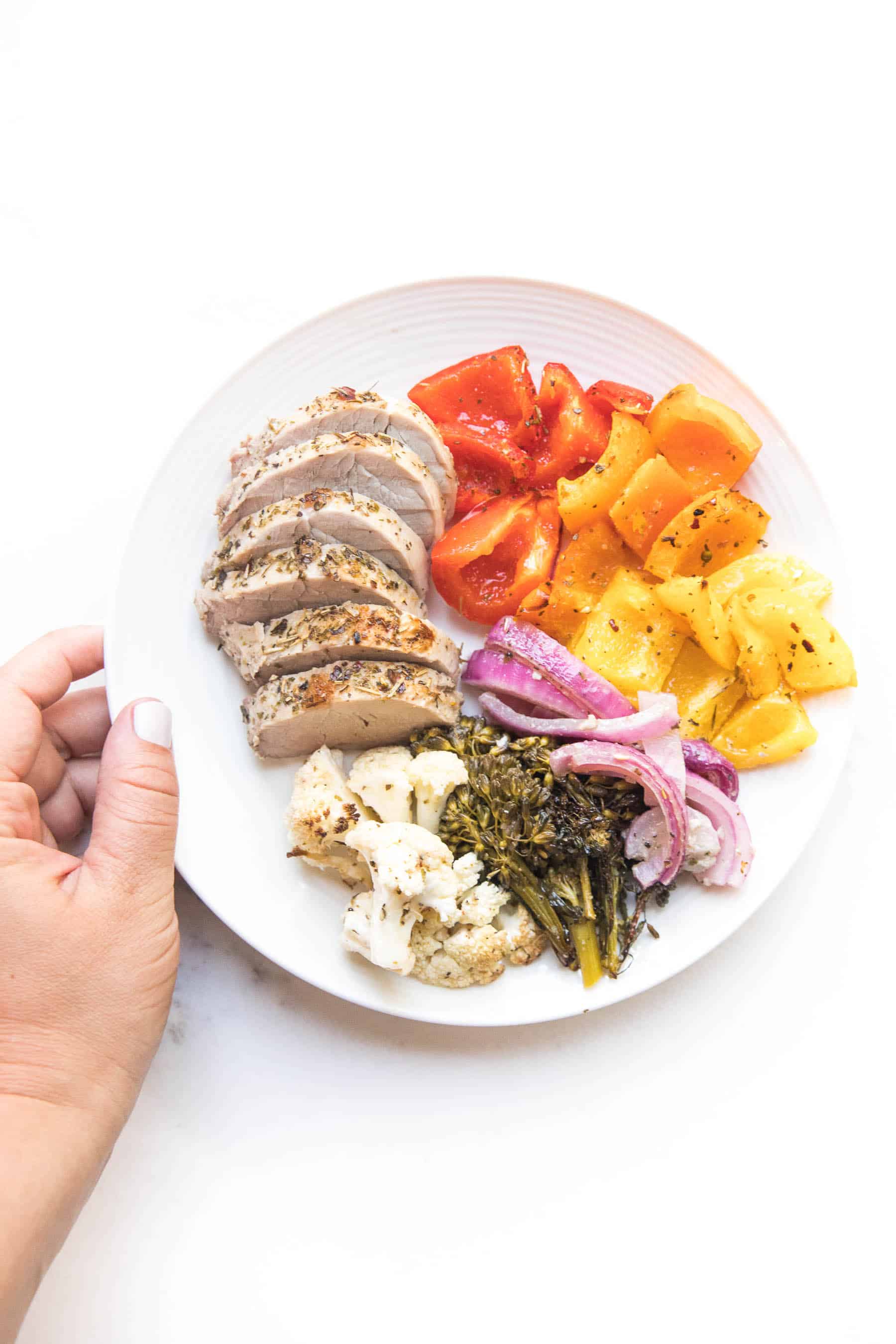 hand holding a white plate with sliced pork tenderloin and rainbow vegetables 