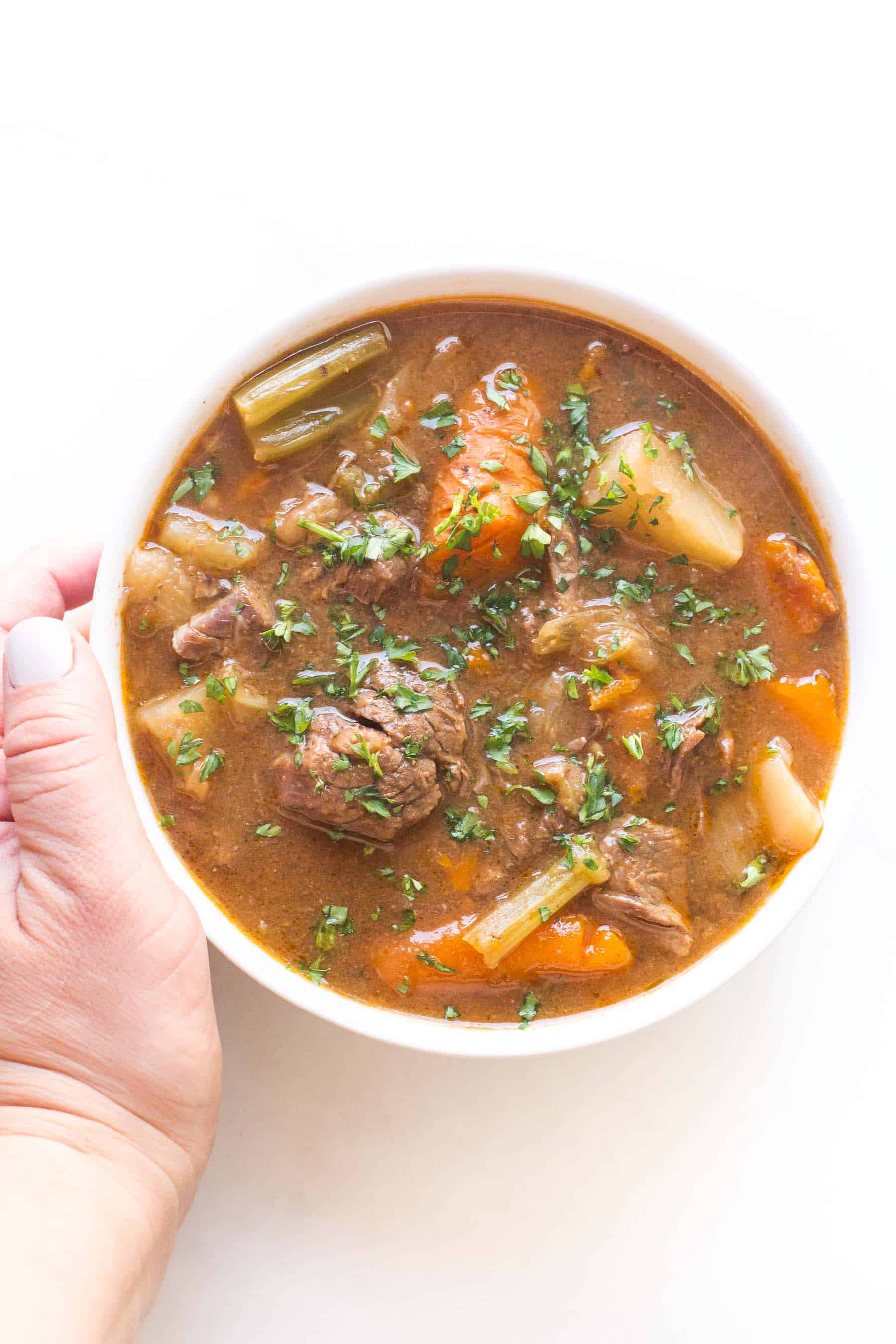 hand holding beef stew in a white bowl on a white background