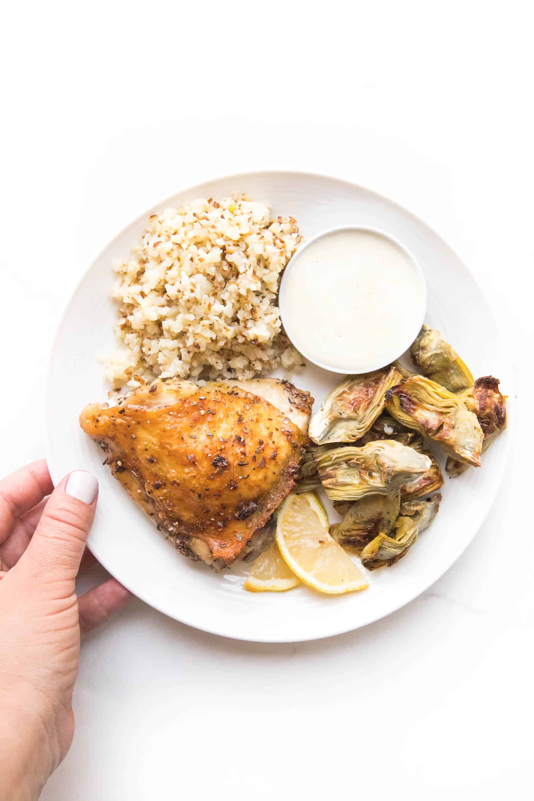 hand holding a white plate with chicken thigh, roasted artichoke hearts and cauliflower rice