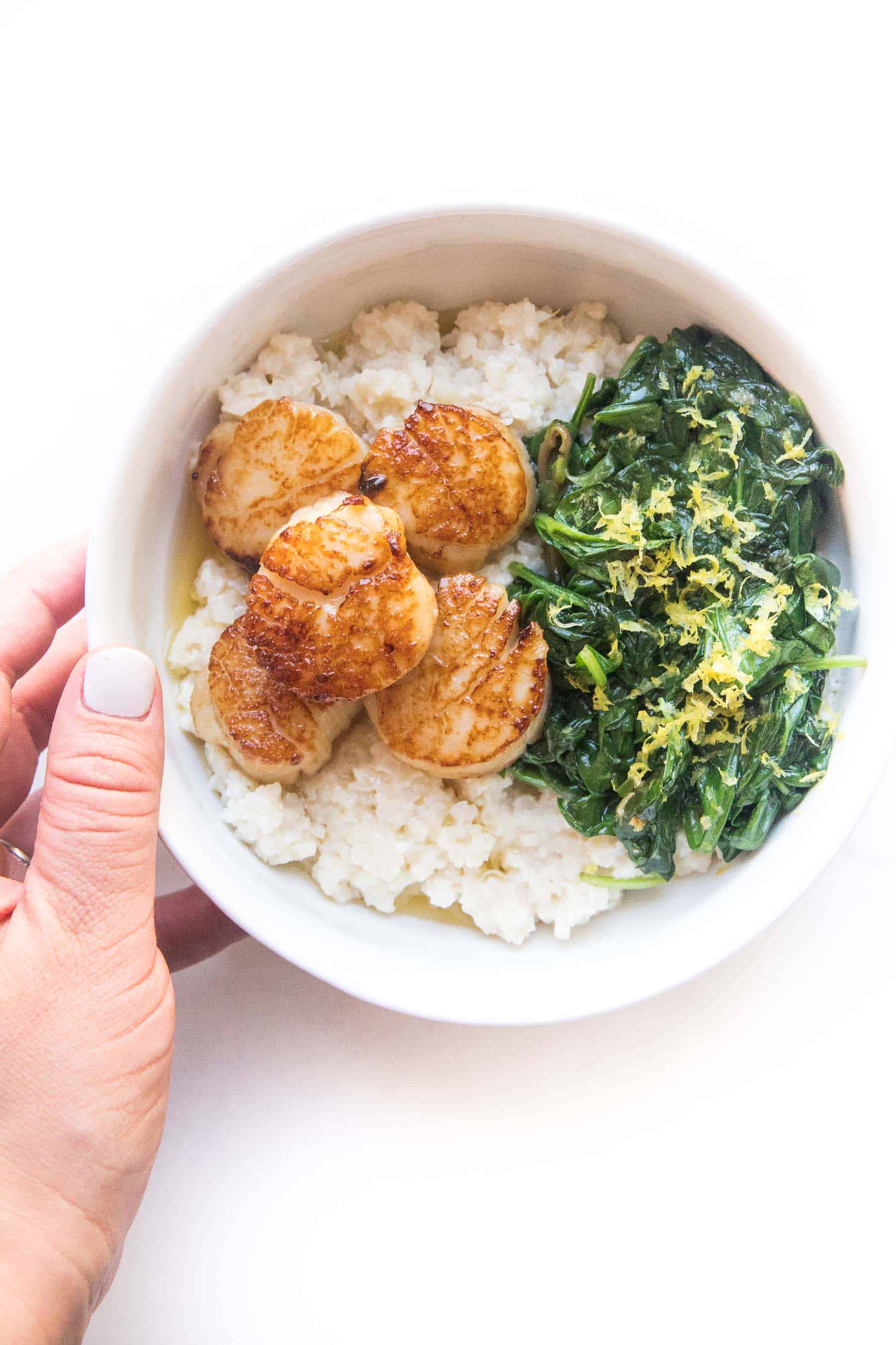 hand holding Seared scallops in a white bowl with cauliflower rice risotto + sauteed spinach