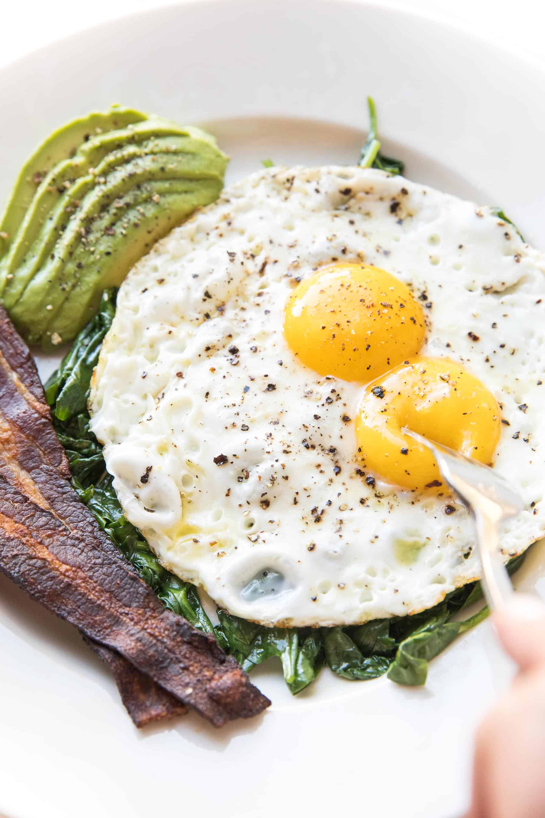 fork breaking sunny side up eggs with sauteed spinach, bacon + avocado